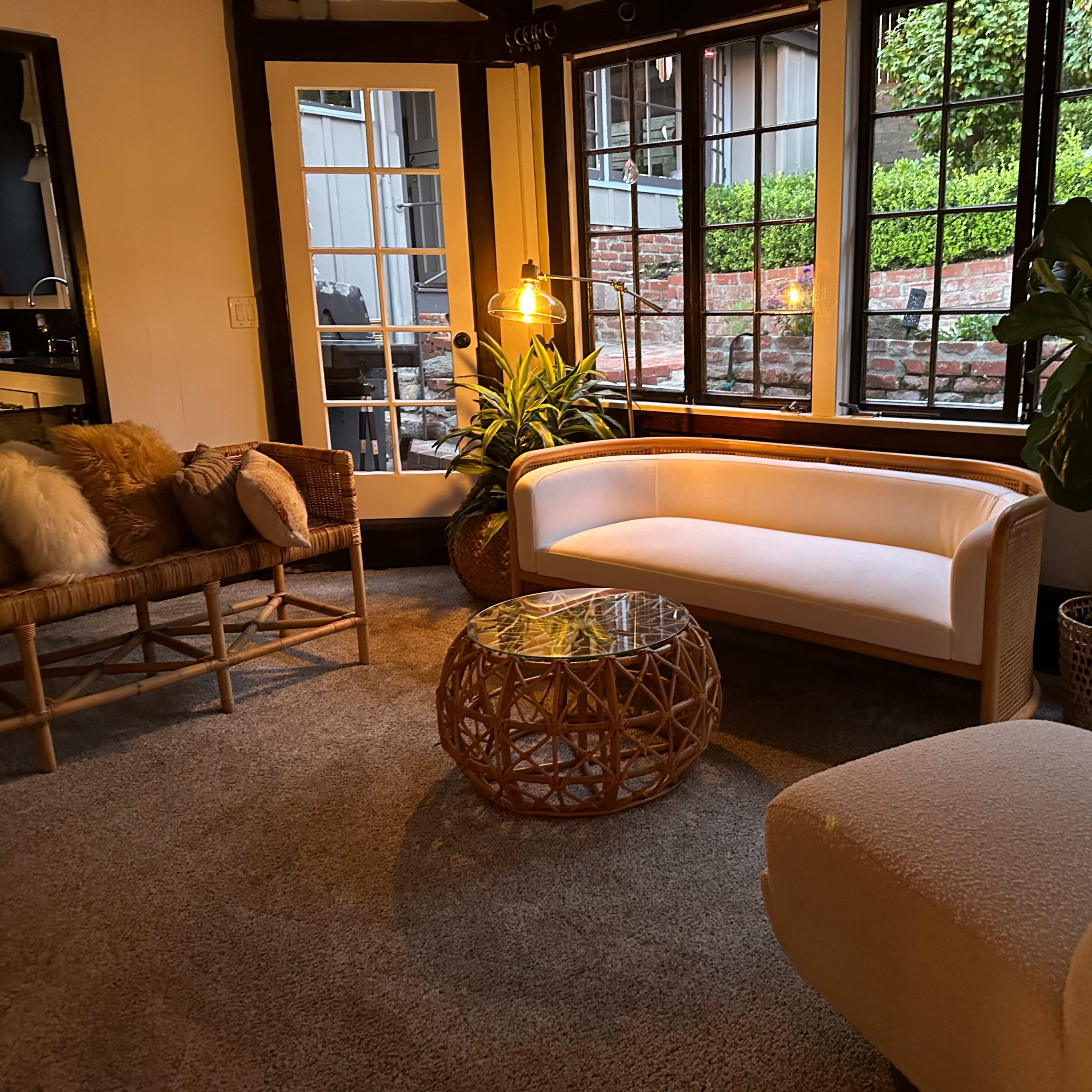 A cozy living room with a light-colored couch, a woven coffee table, and large windows allowing natural light to illuminate the space.