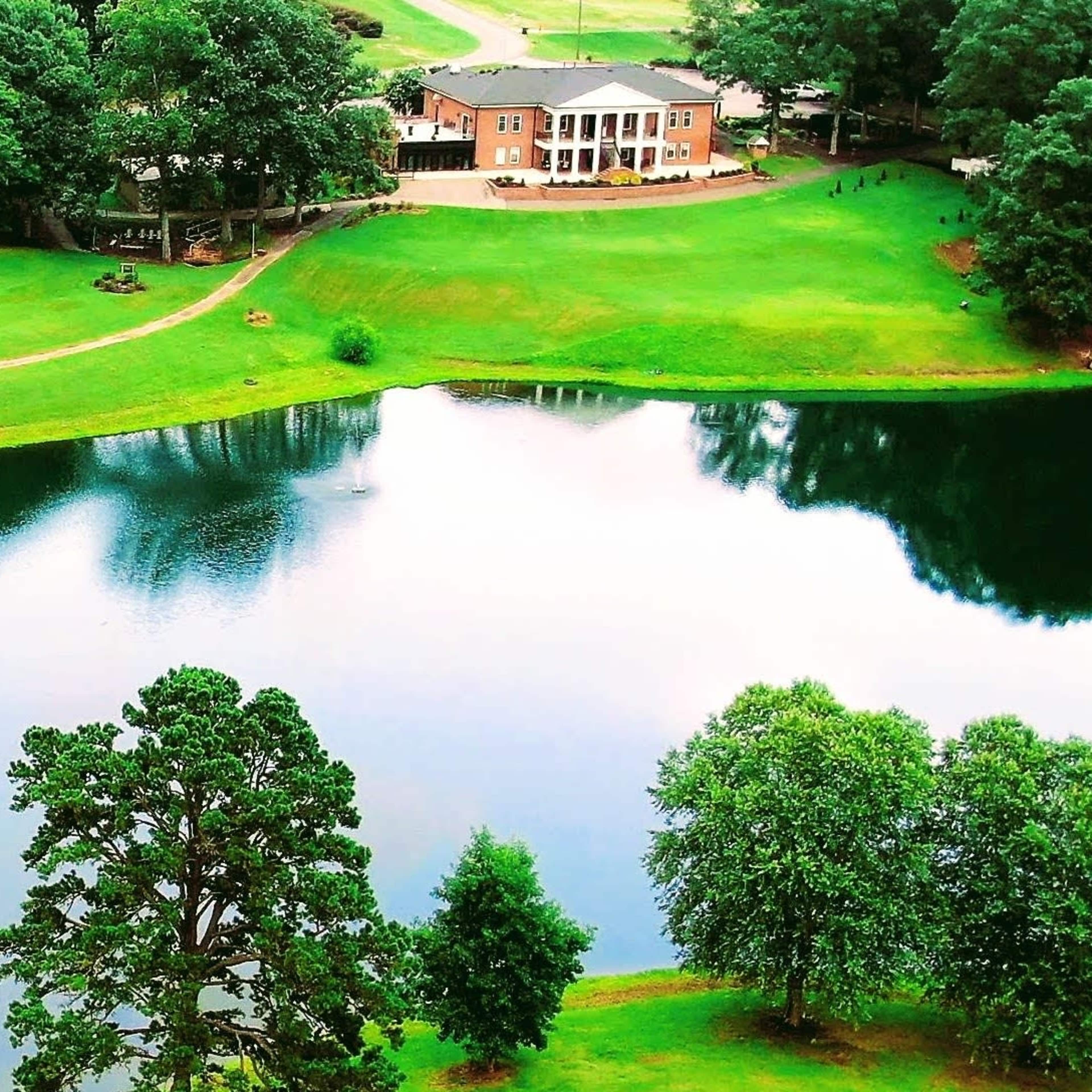 A clear lake reflects a lush green landscape, with a building and trees visible along the water's edge.