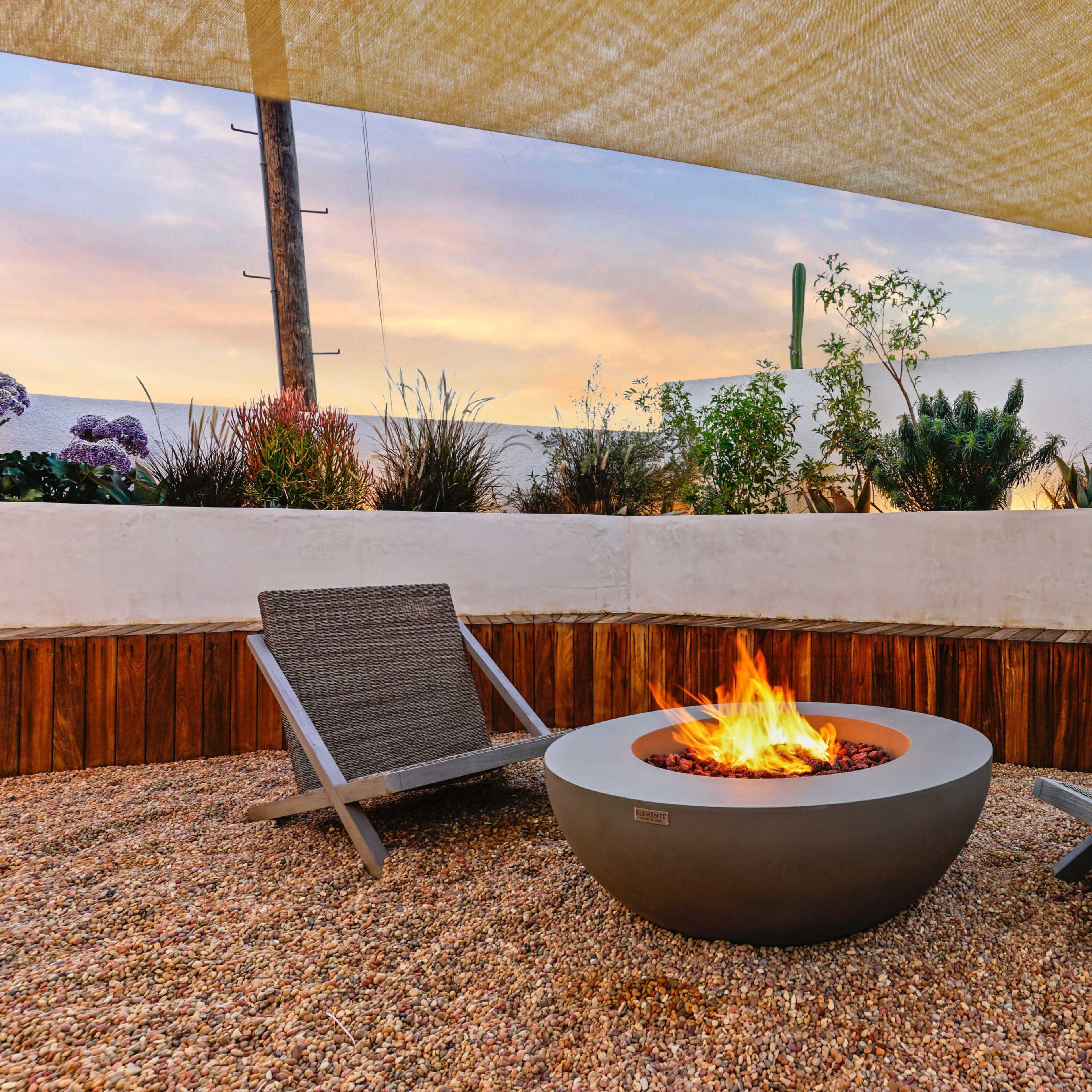Two lounge chairs positioned around a circular fire pit on a gravel surface, with a backdrop of a garden and a sunset sky partially covered by a shade structure.