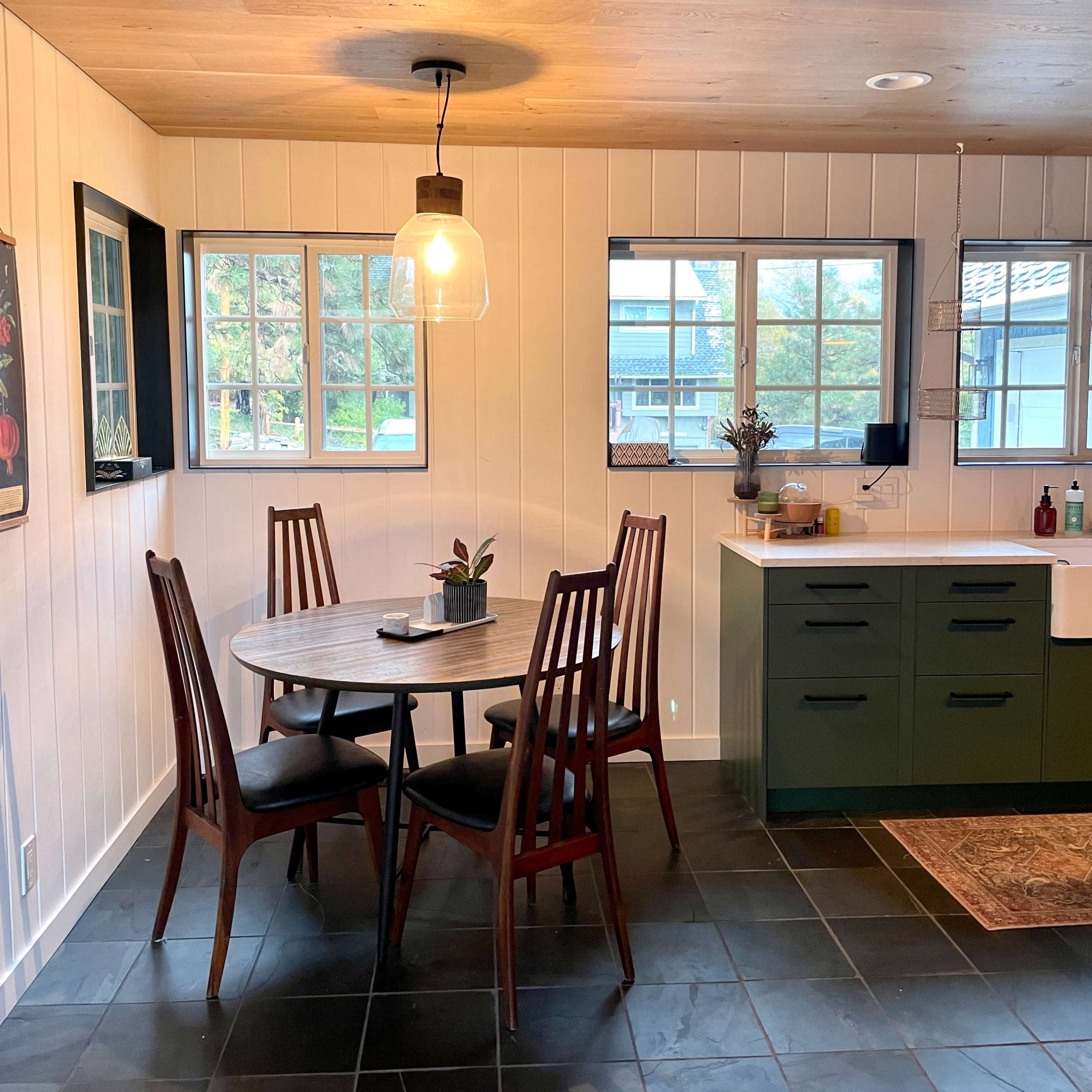A kitchen with a round dining table surrounded by four chairs, large windows providing natural light, and a green cabinetry setup.