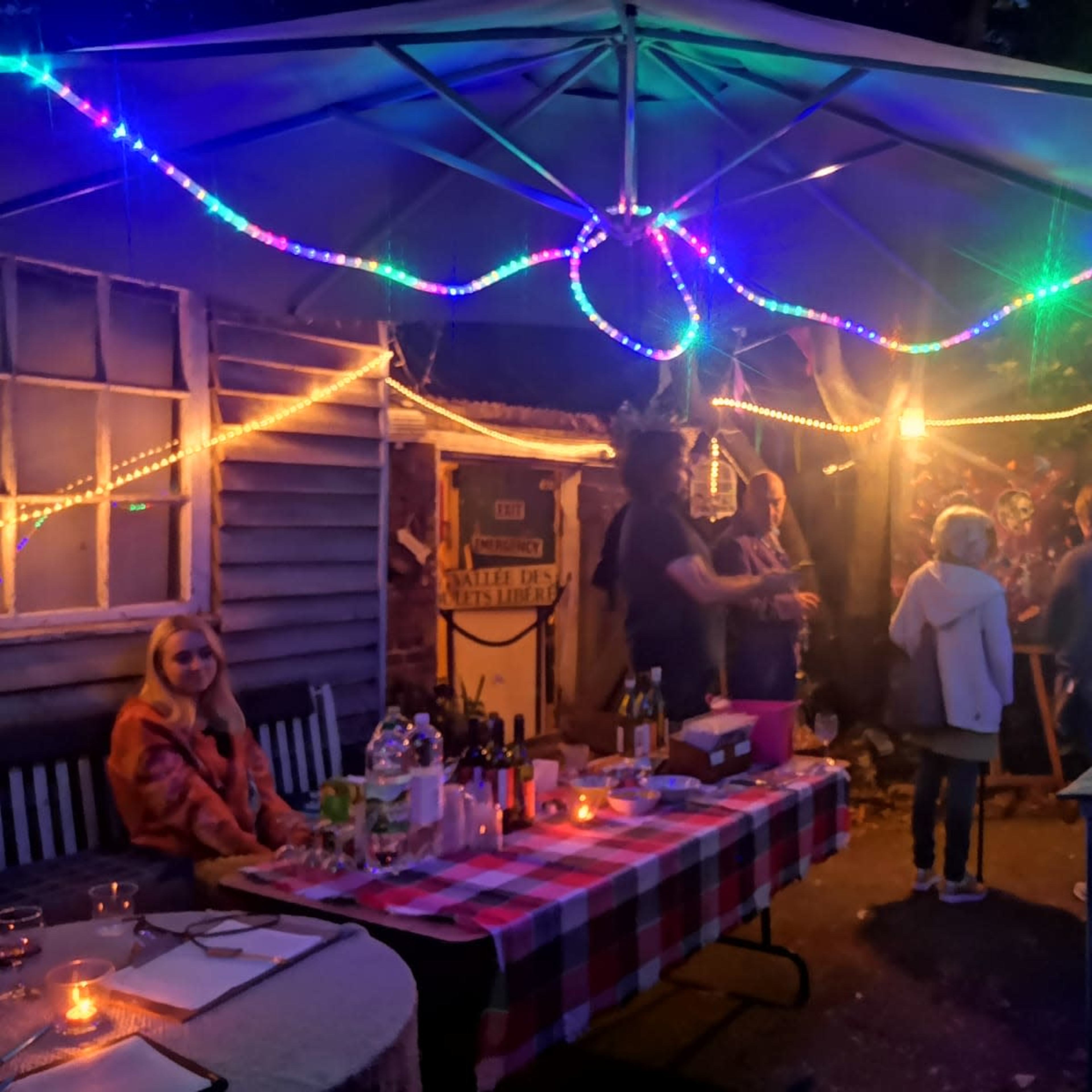 A group of people gathers under a lit tent adorned with colorful lights in a backyard setting at night.