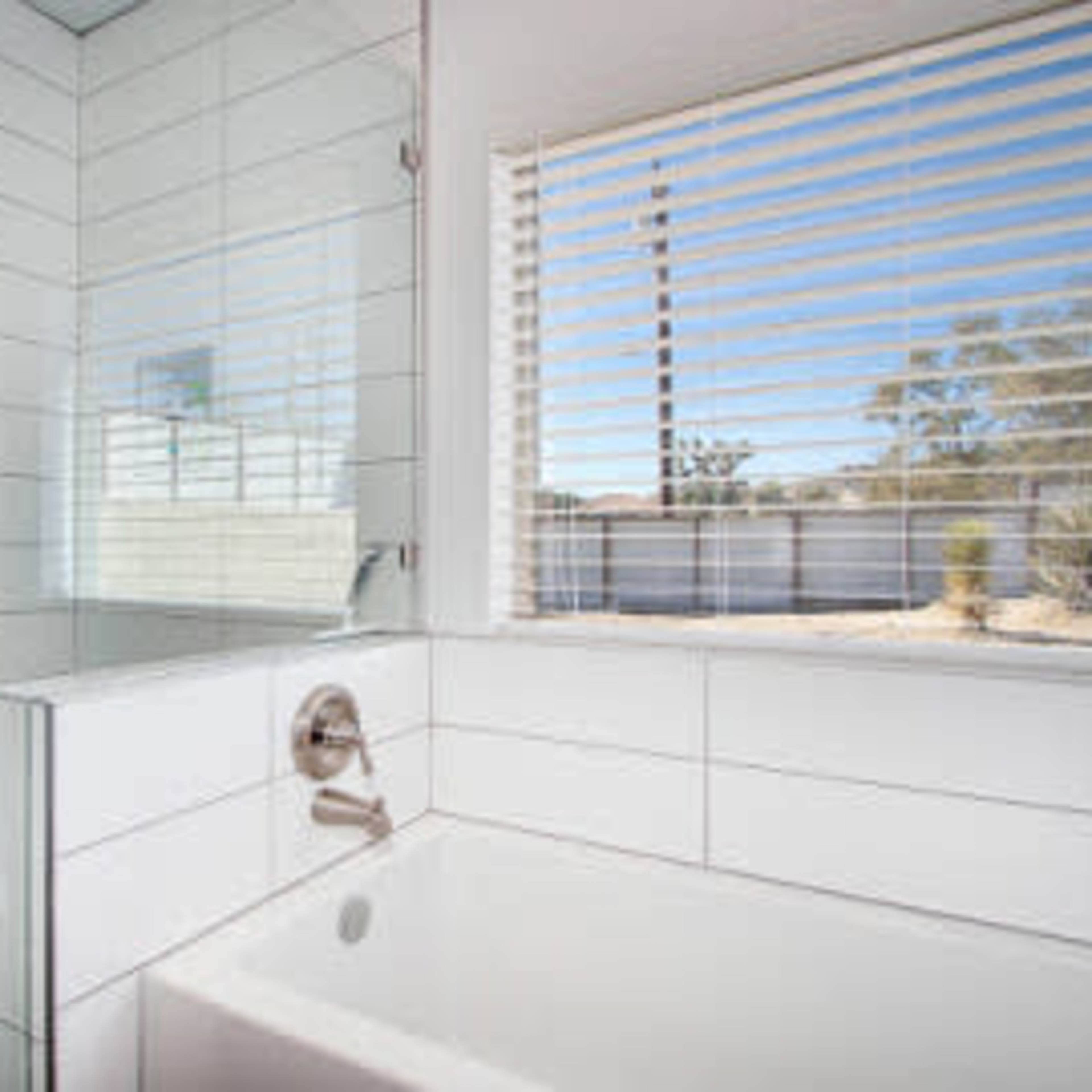 A modern bathroom featuring a glass shower, a white bathtub, and a window with horizontal blinds overlooking a desert landscape.