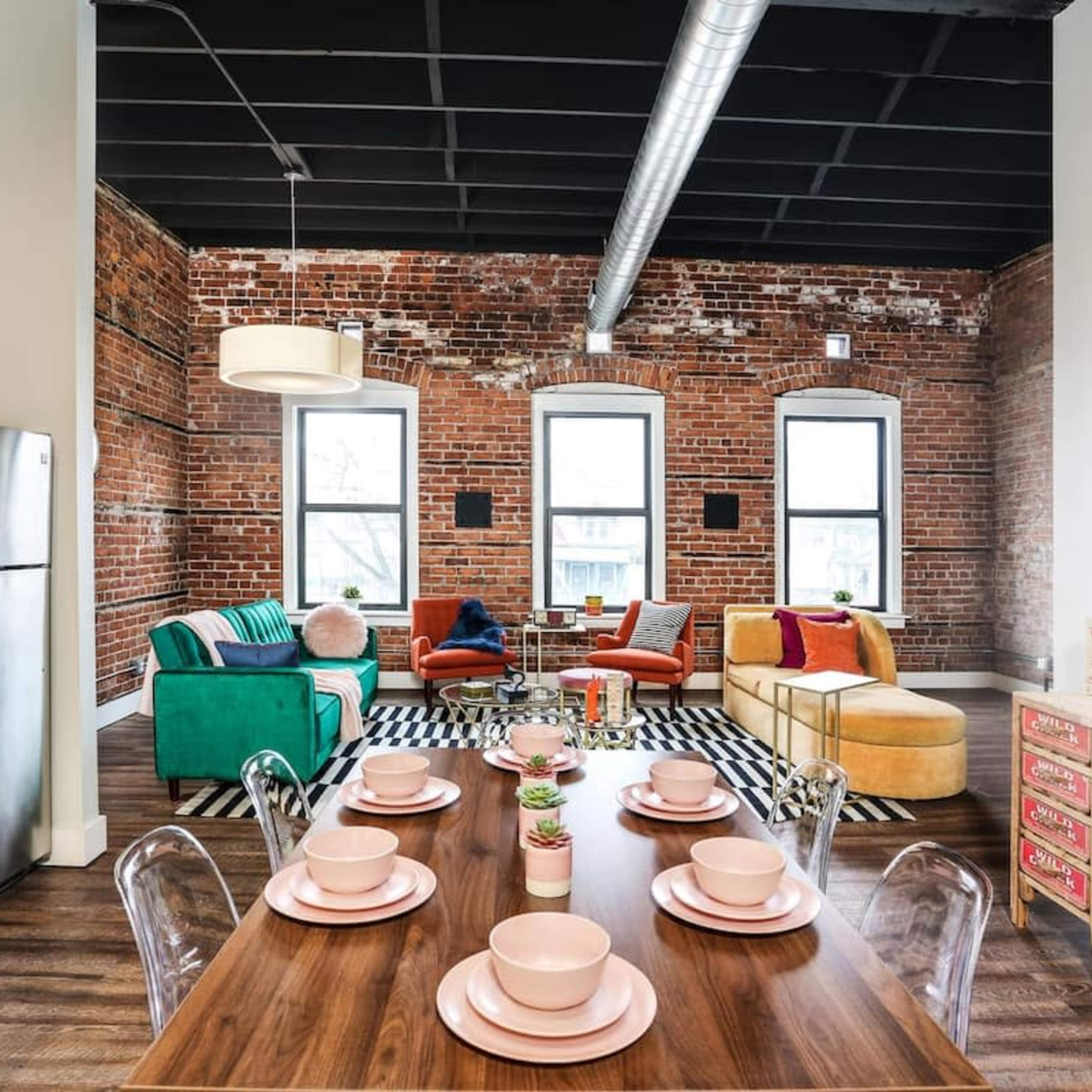 A modern living space featuring exposed brick walls, large windows, a mix of colorful furniture, and a dining table set with pink plates.