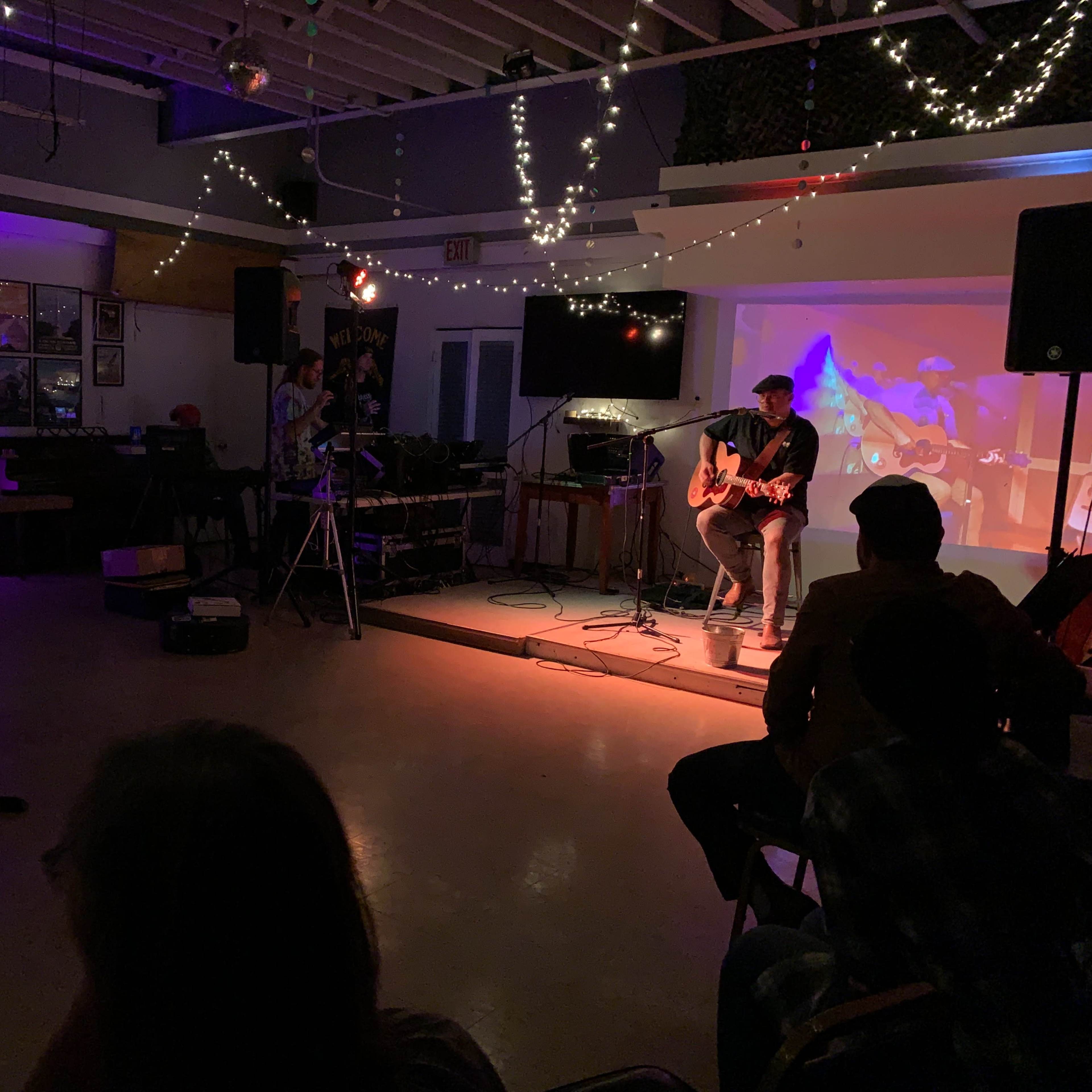 A musician plays guitar on stage while a small audience observes in a dimly lit venue adorned with string lights.