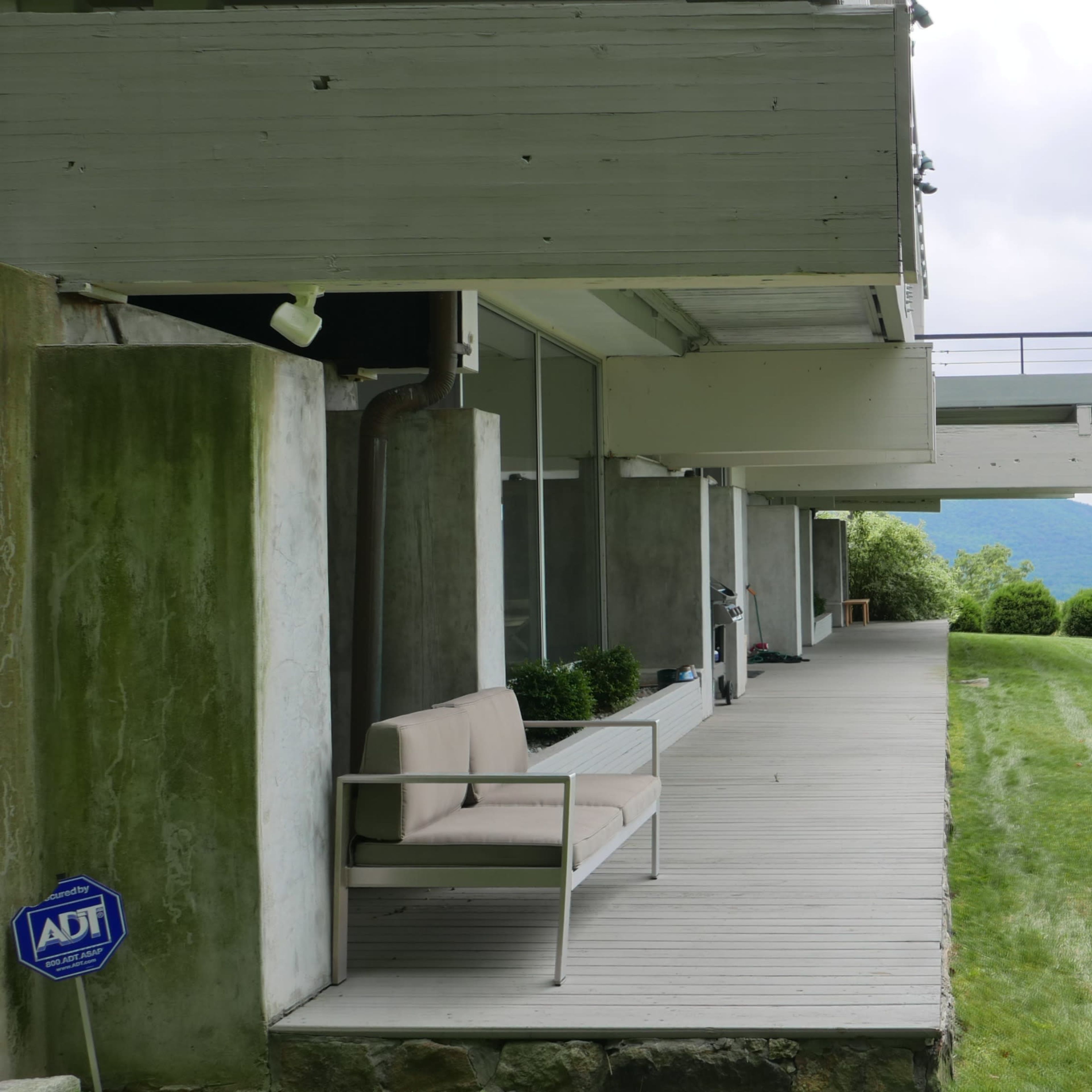 A modern building with a wooden deck and a gray sofa, overlooking a lawn and distant mountains under a cloudy sky.