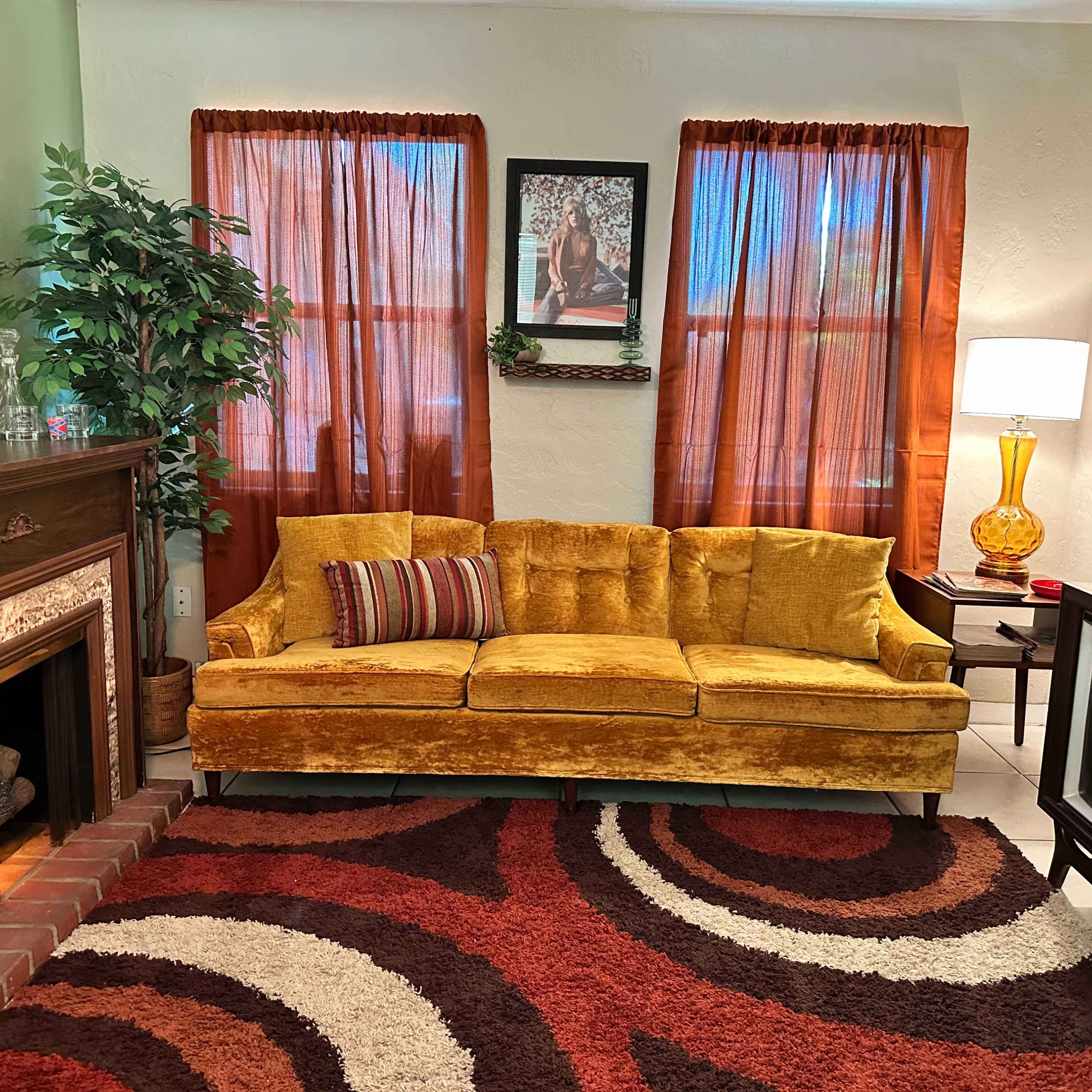 A living room with a yellow velvet couch, a vintage television, a patterned rug, and orange curtains.