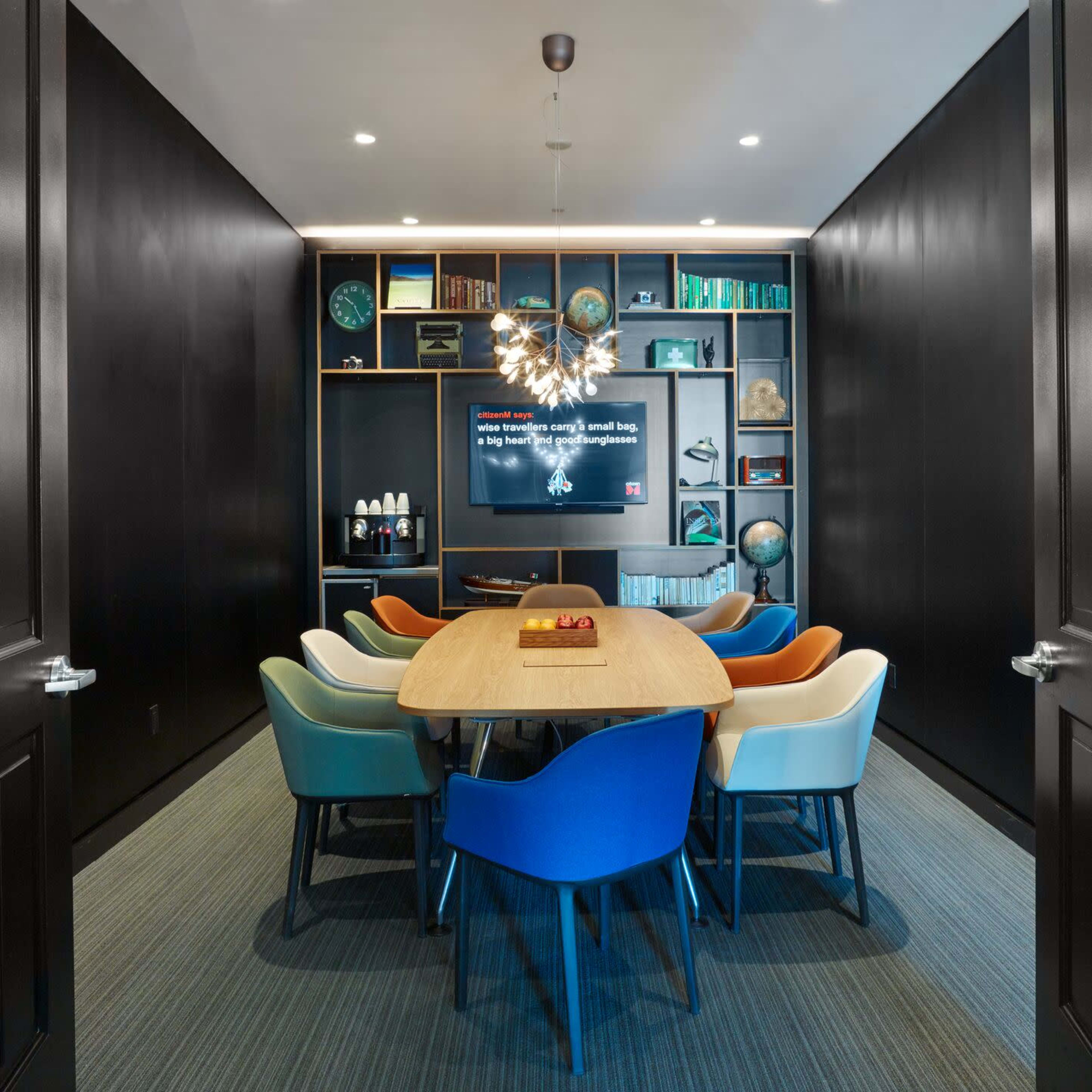 A modern conference room features a large wooden table surrounded by colorful chairs and a television mounted on a black wall, with shelves displaying various items in the background.