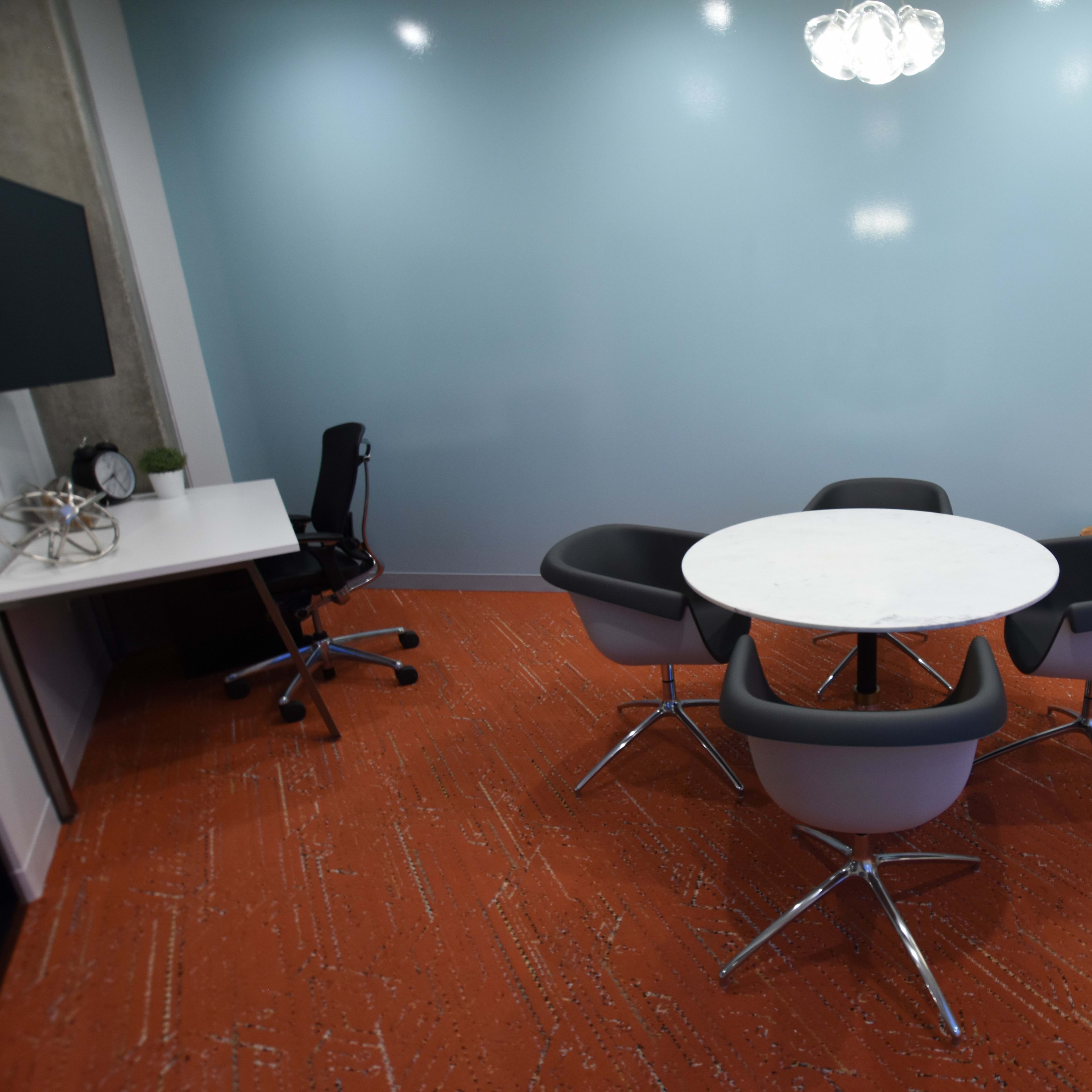 A small conference room features a round table surrounded by four chairs, with a lamp and a desk in the background.