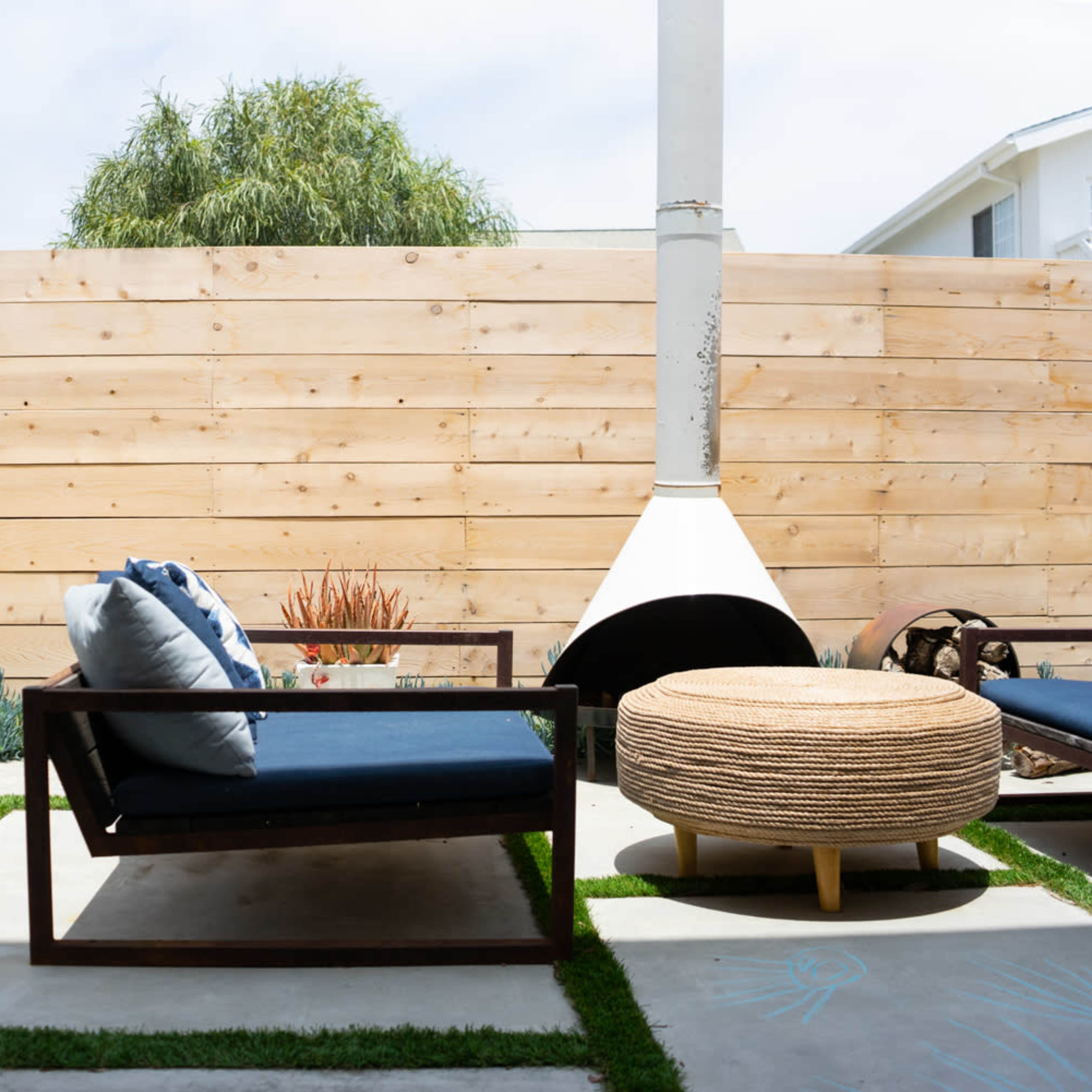 A patio area with a wooden fence, a lounge chair with blue cushions, a round wicker table, and patches of grass and concrete flooring.