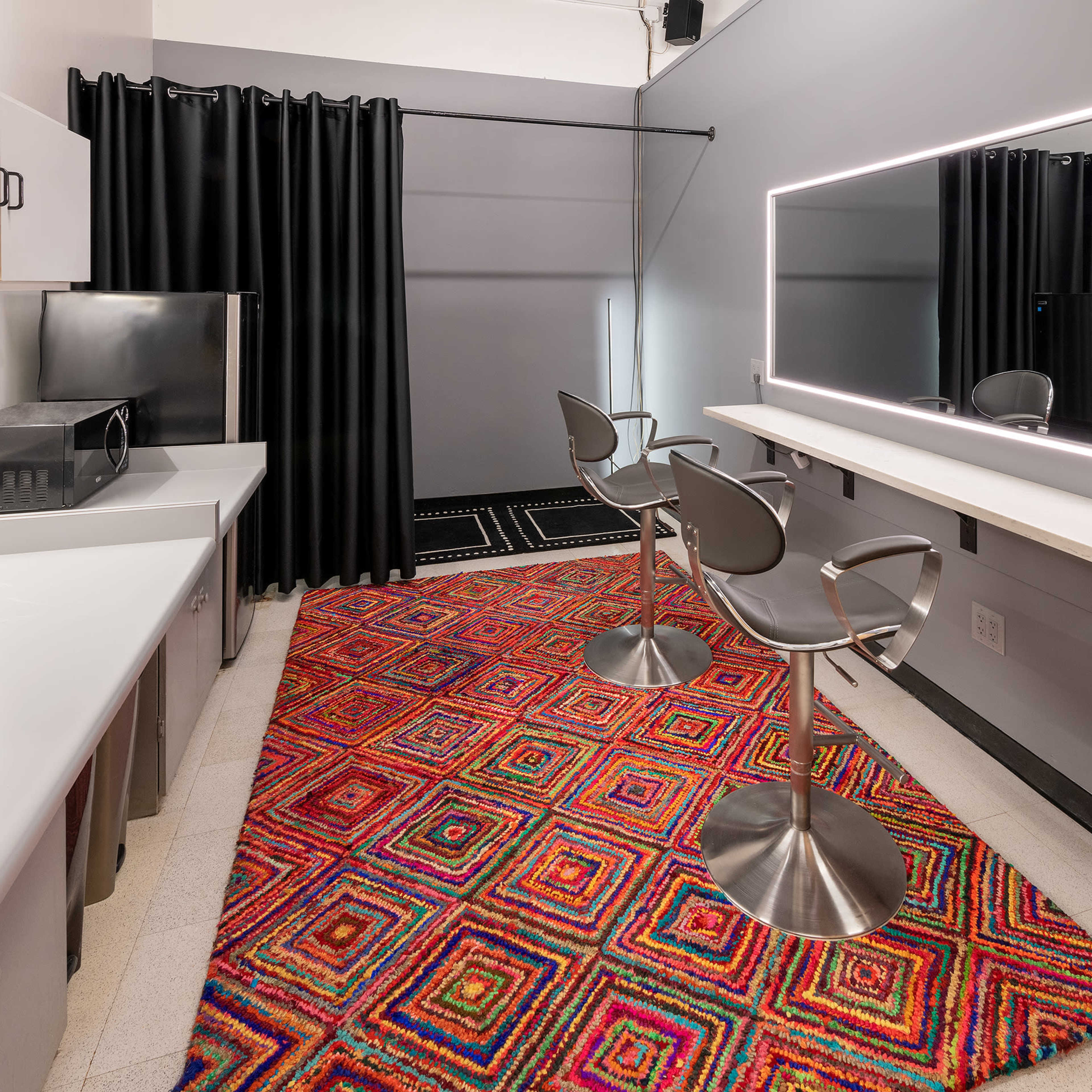 A modern makeup room featuring two high stools, a backlit mirror, and a colorful geometric rug on the floor.