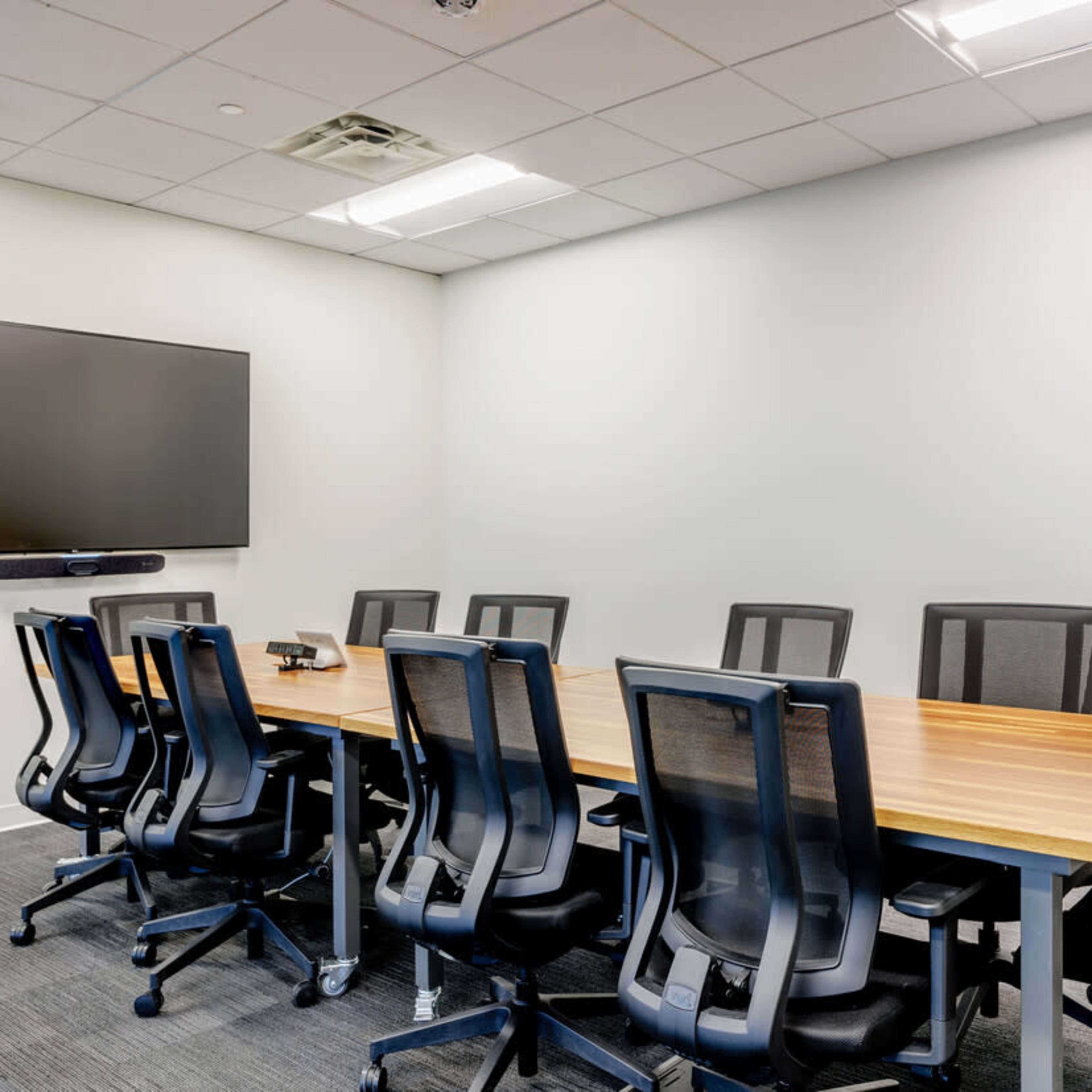 A sparsely furnished meeting room features a long wooden table surrounded by black office chairs and a mounted television on the wall.