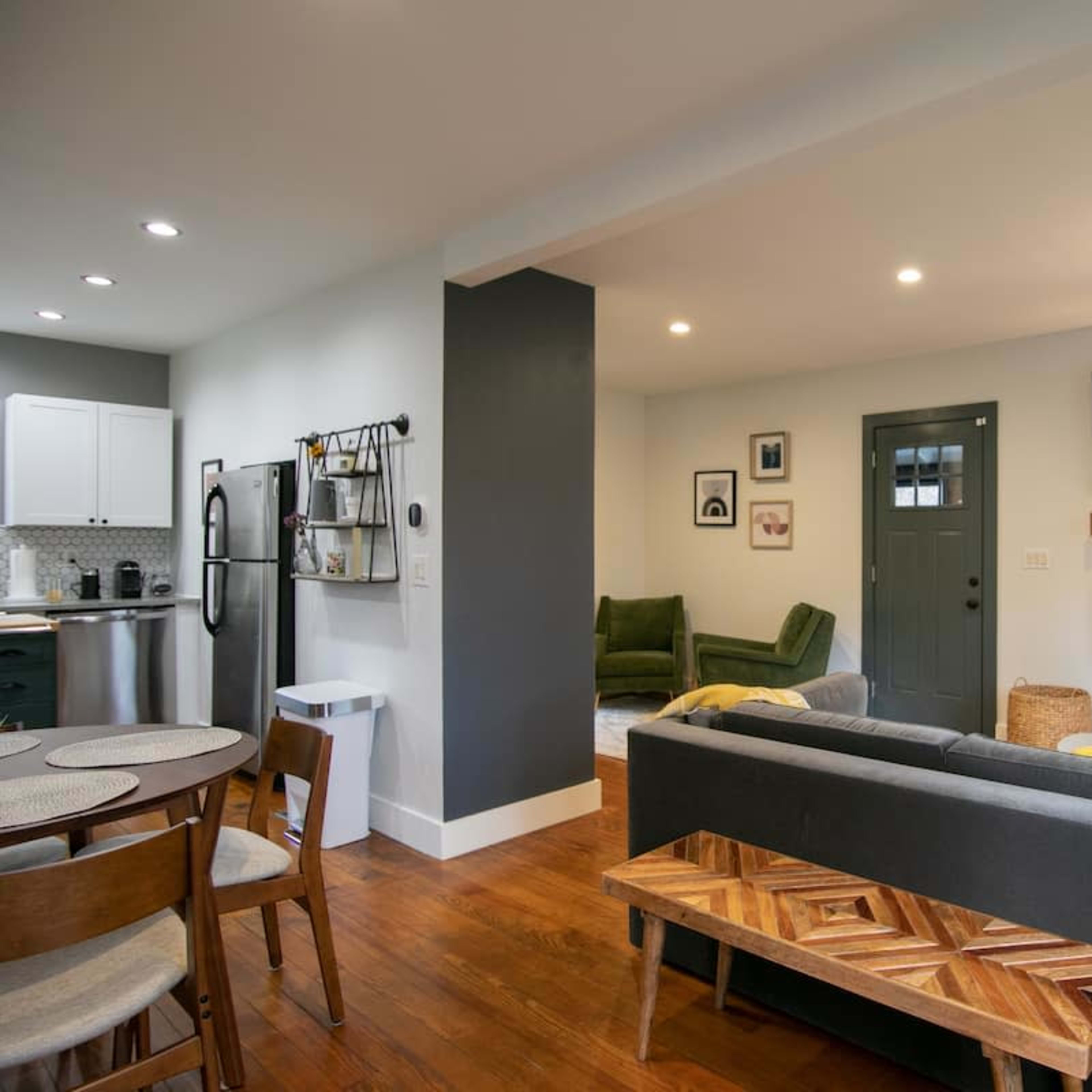 A modern living room with a gray sofa, a wooden coffee table, and a mounted TV on the wall, complemented by a green armchair and a doorway leading outside.
