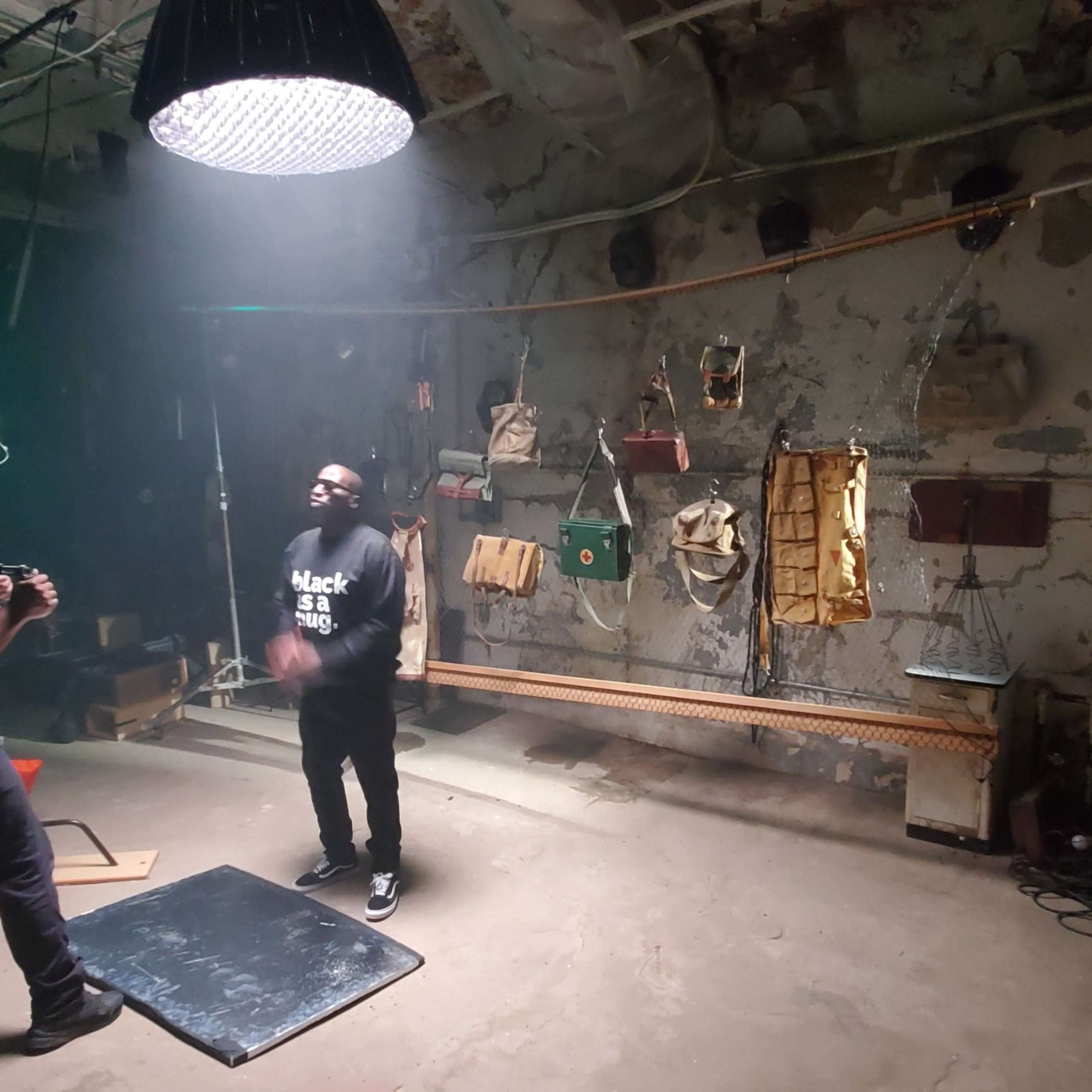 A person stands on a mat in a dimly lit industrial space while another person operates a camera nearby.