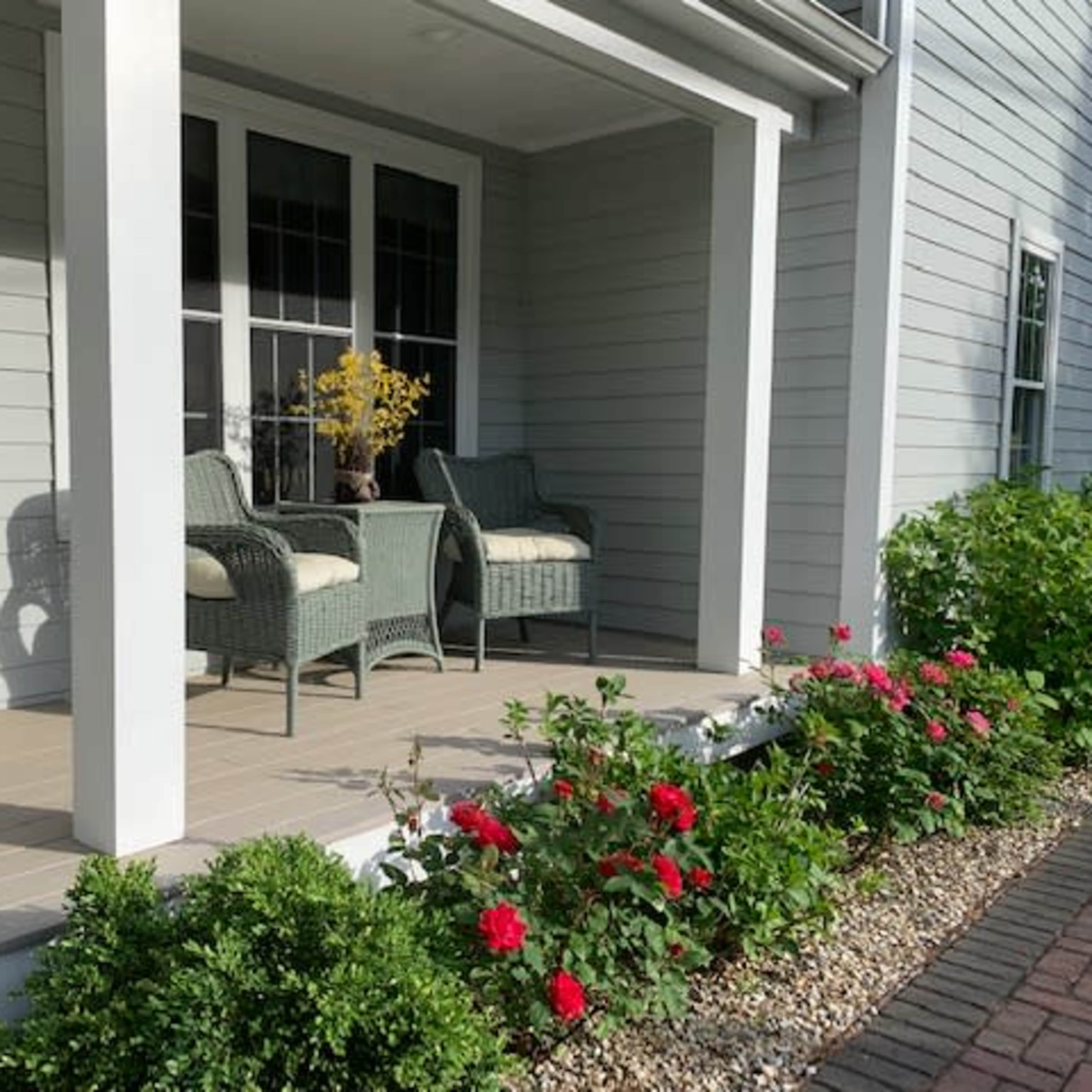 A house porch featuring two wicker chairs, potted plants, and a row of blooming flowers along a gravel border.