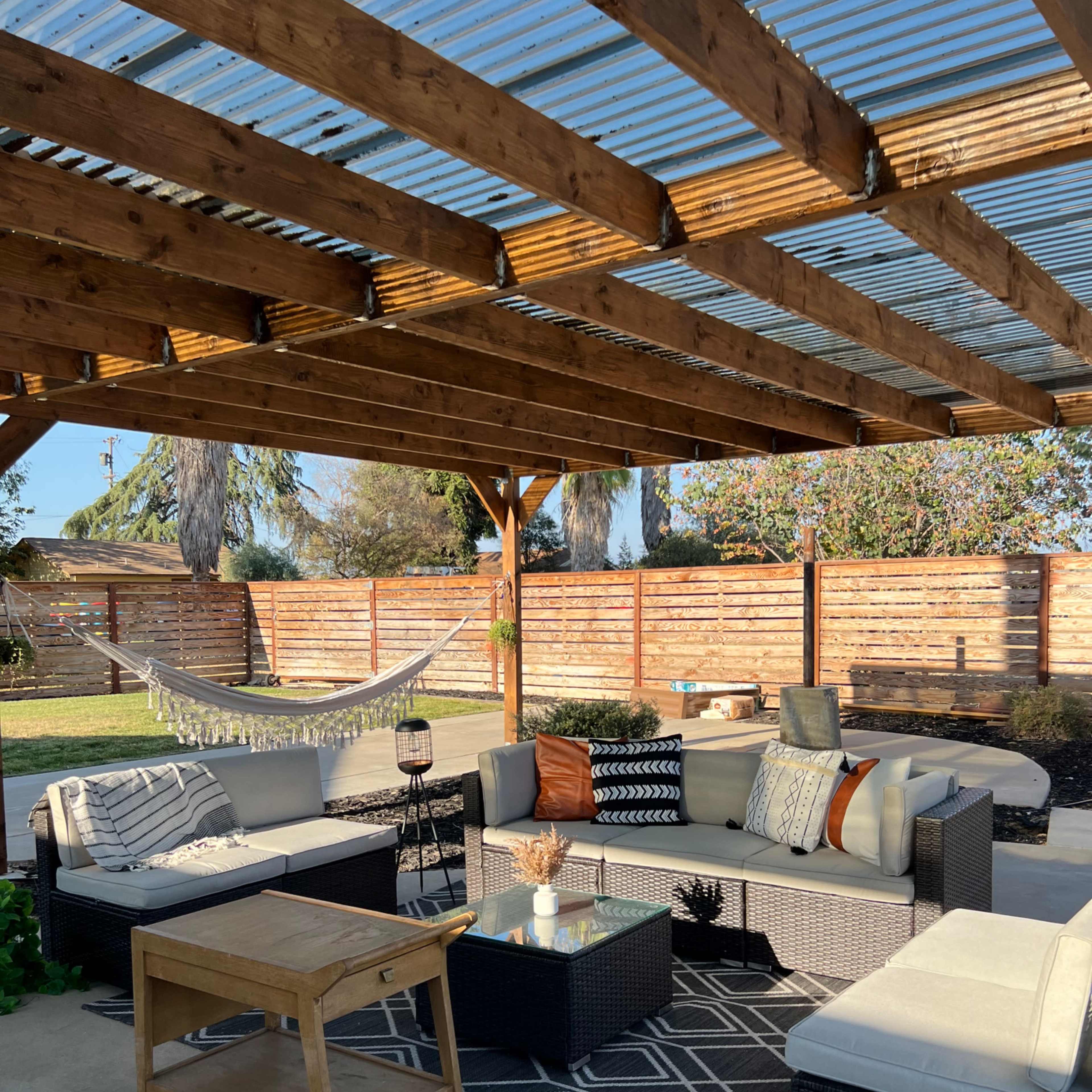 An outdoor patio is arranged with a sectional sofa, coffee table, and a hammock beneath a wooden pergola.