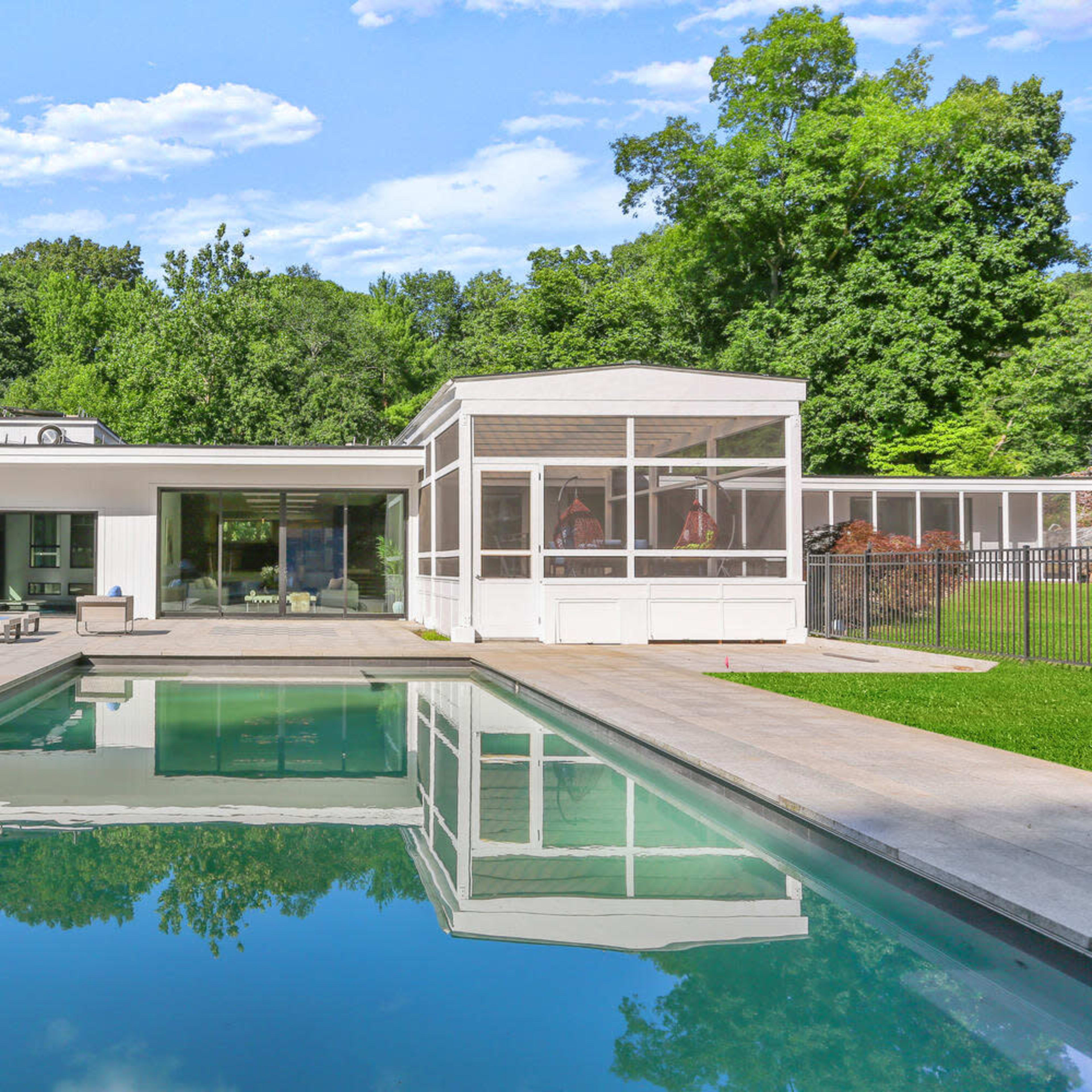 A modern house with large windows overlooks a rectangular swimming pool surrounded by a patio and green landscape.