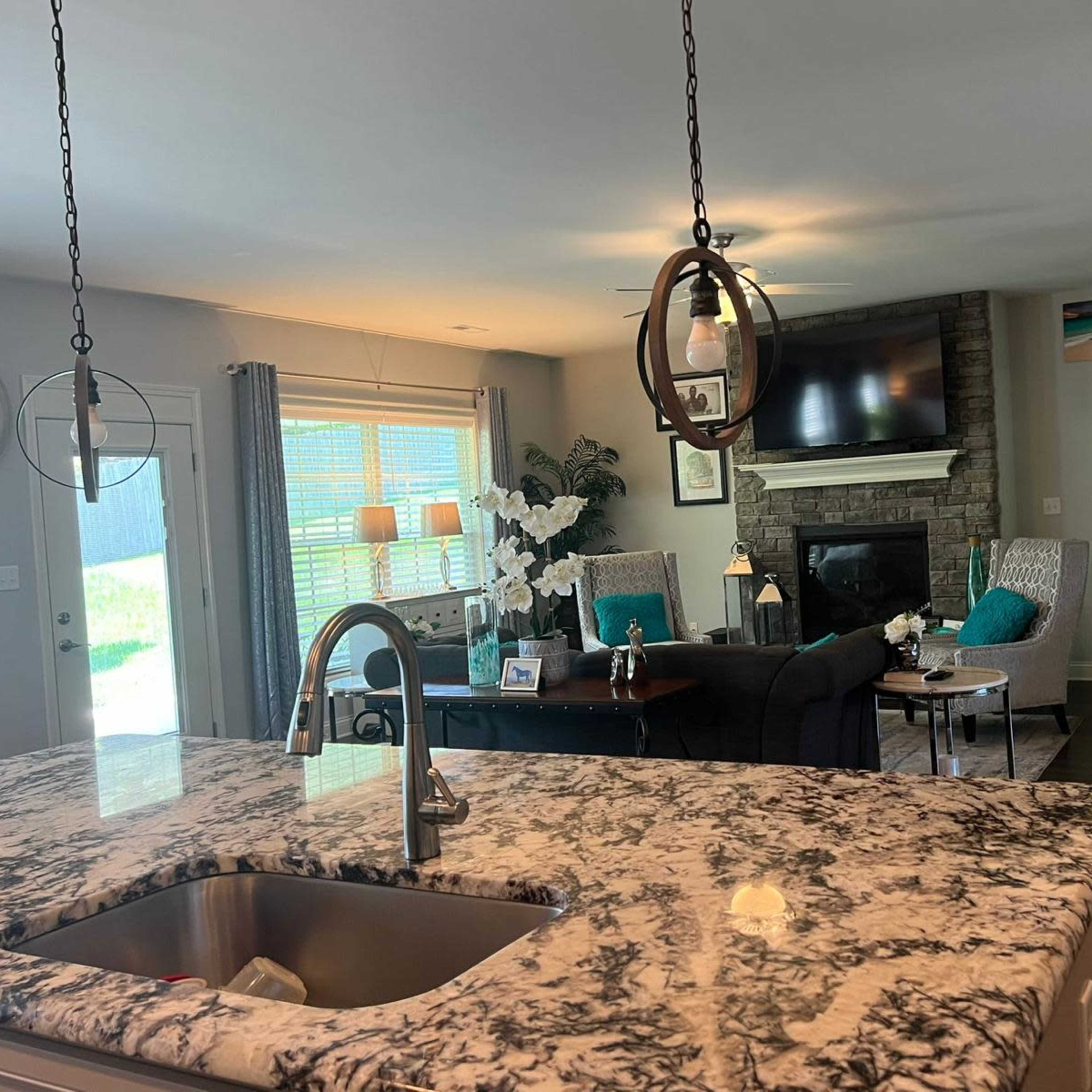 A living room with a stone fireplace and large windows, viewed from a kitchen with a granite countertop.
