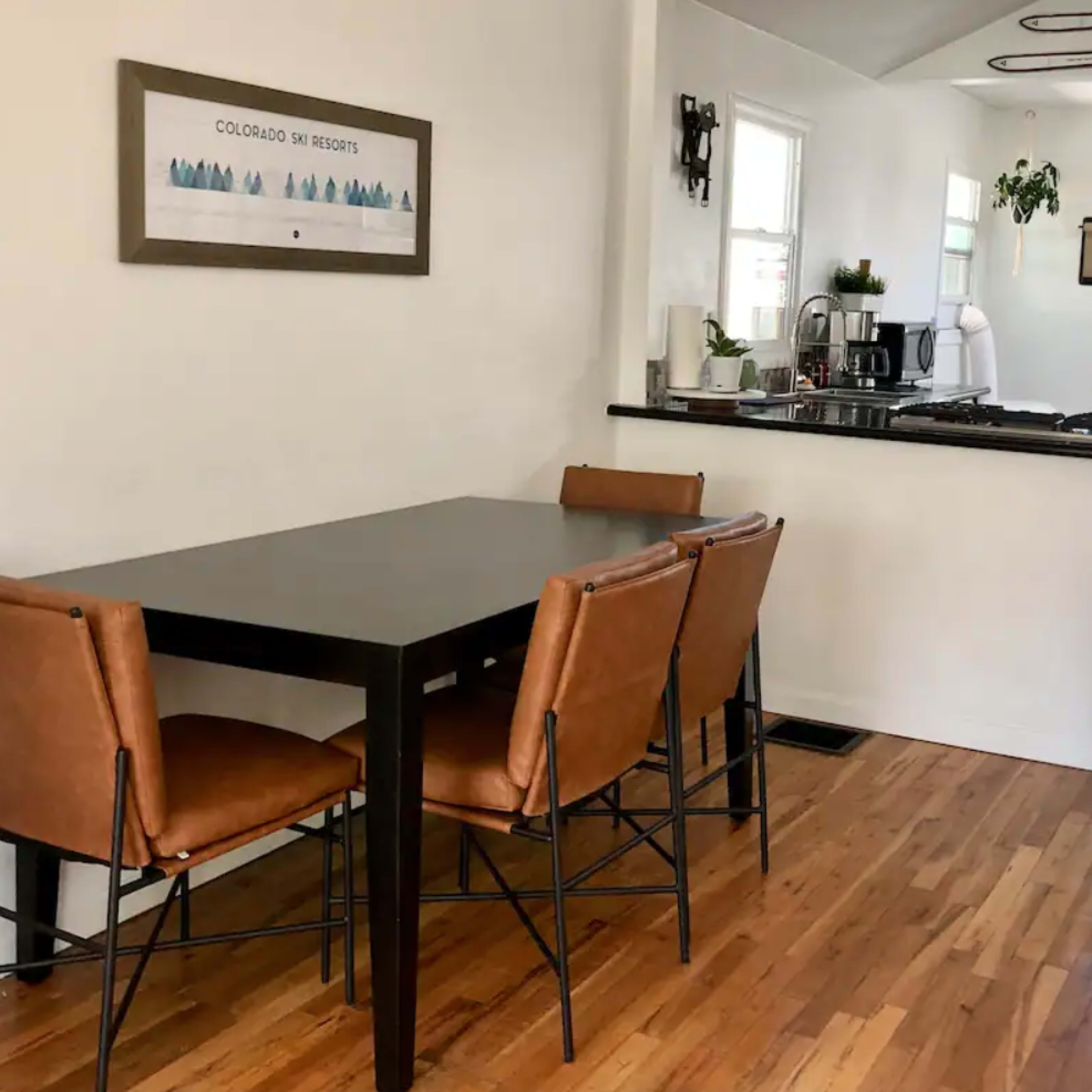 A simple dining area features a rectangular table with four brown chairs, adjacent to a kitchen with modern appliances and a doorway leading outside.