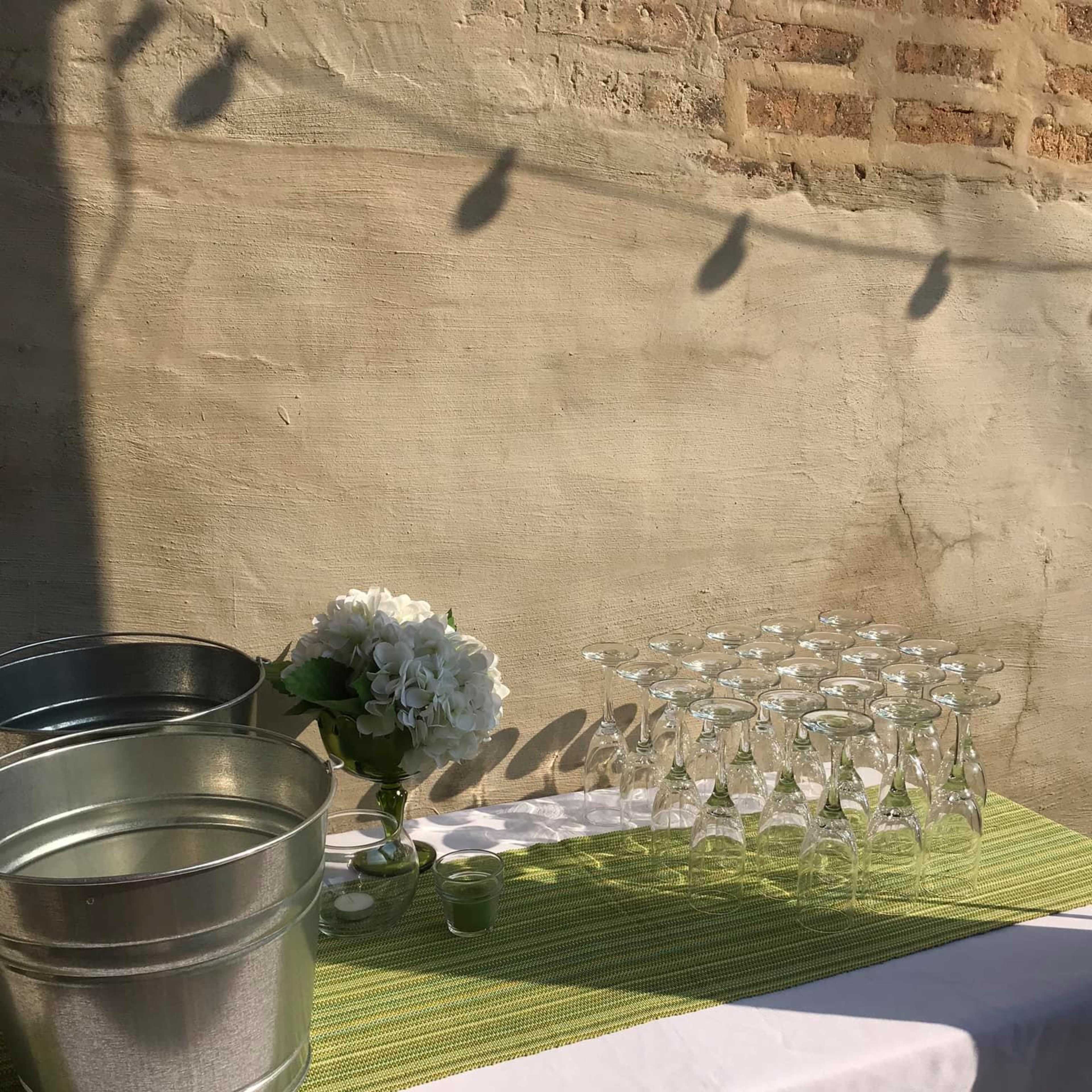 A table setup featuring two metal buckets, a floral arrangement, and a row of clear glassware on a green-striped tablecloth against a textured wall.