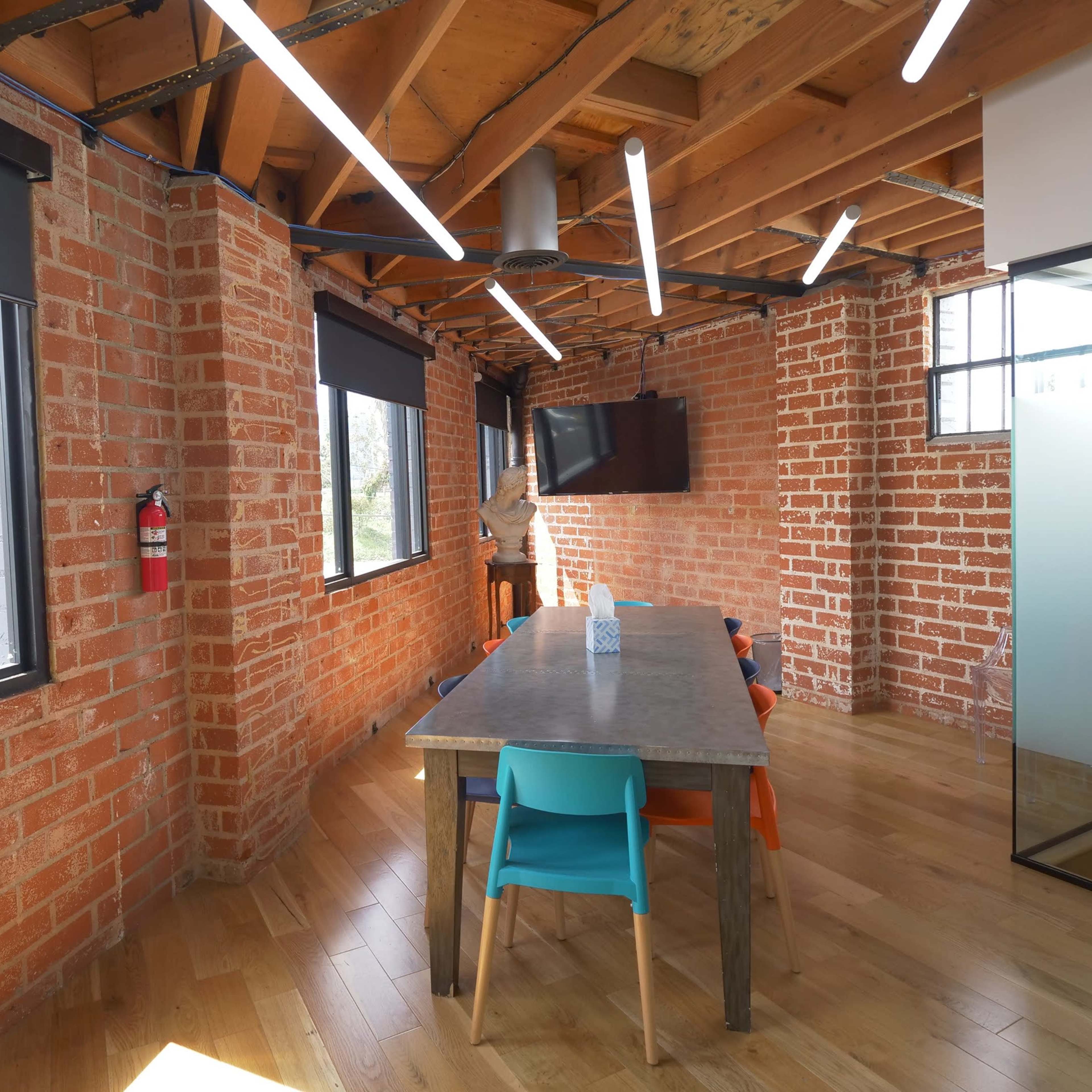 A modern office space with exposed brick walls, a wooden table surrounded by colorful chairs, and large windows allowing natural light to enter.