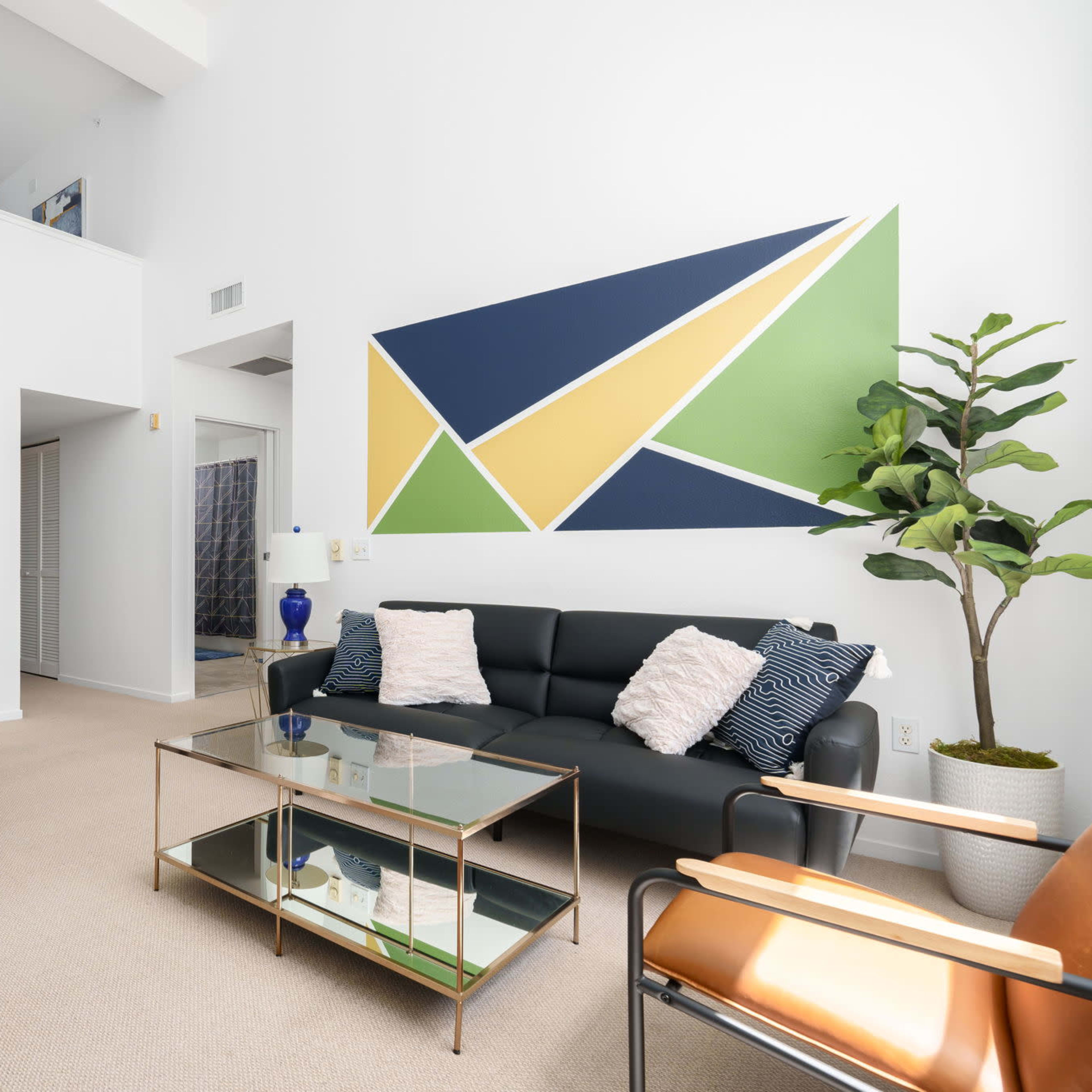 A modern living room featuring a black sofa, a glass coffee table, a potted plant, and a colorful geometric wall mural, with a staircase visible on the left.