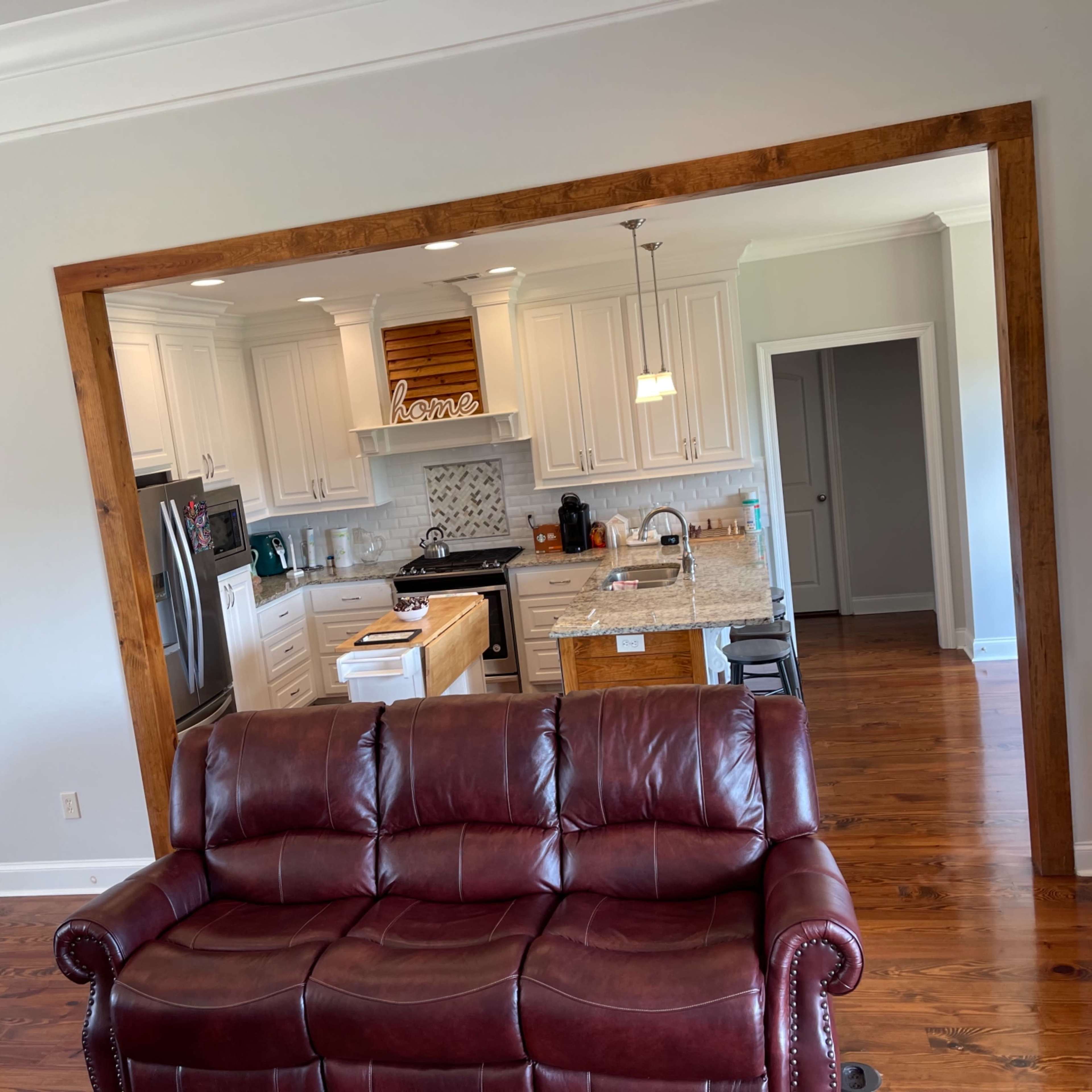 A living room with a brown leather sofa facing an open kitchen area featuring white cabinets and a granite countertop.