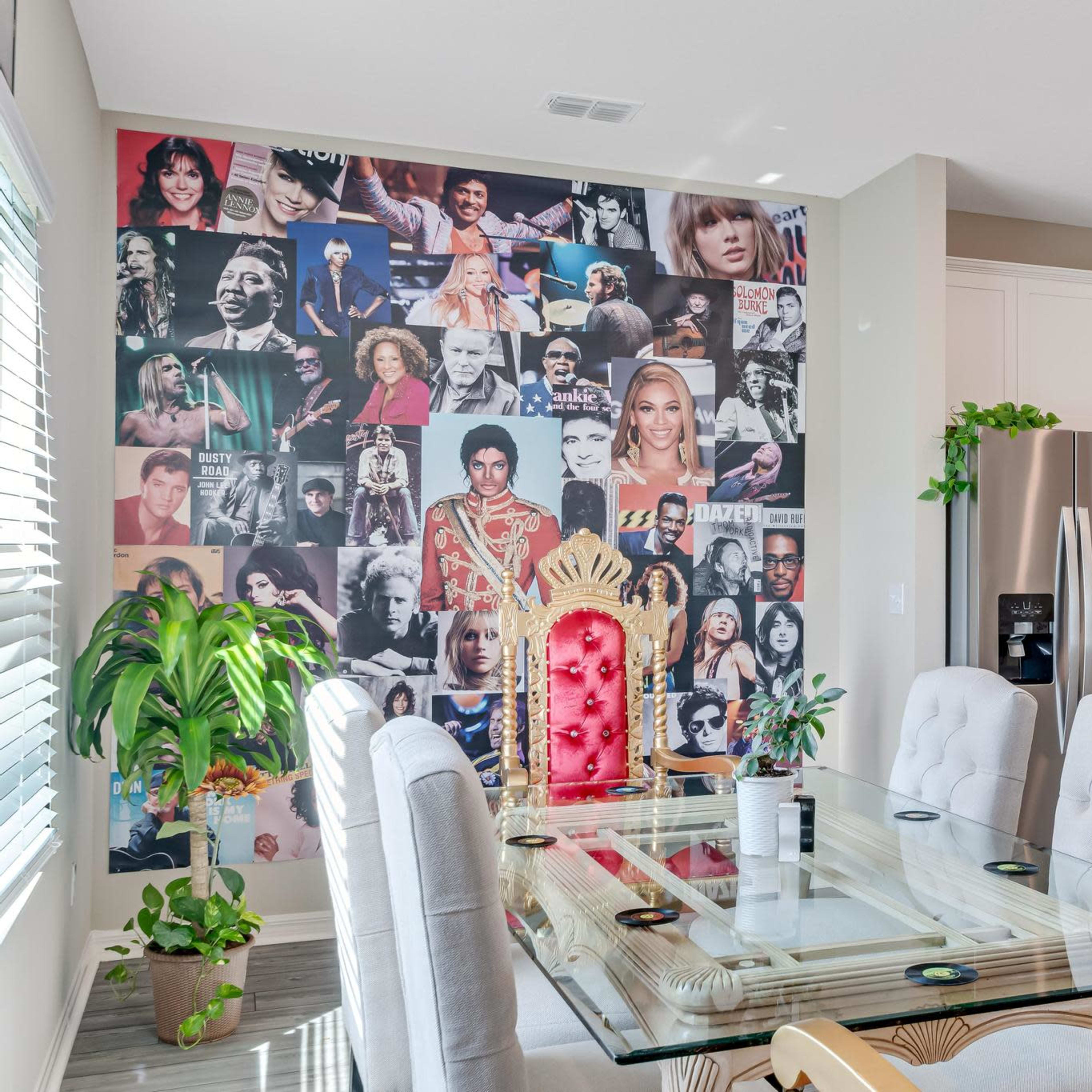 A dining area with a glass table and ornate golden chairs, featuring a wall collage of various celebrities.