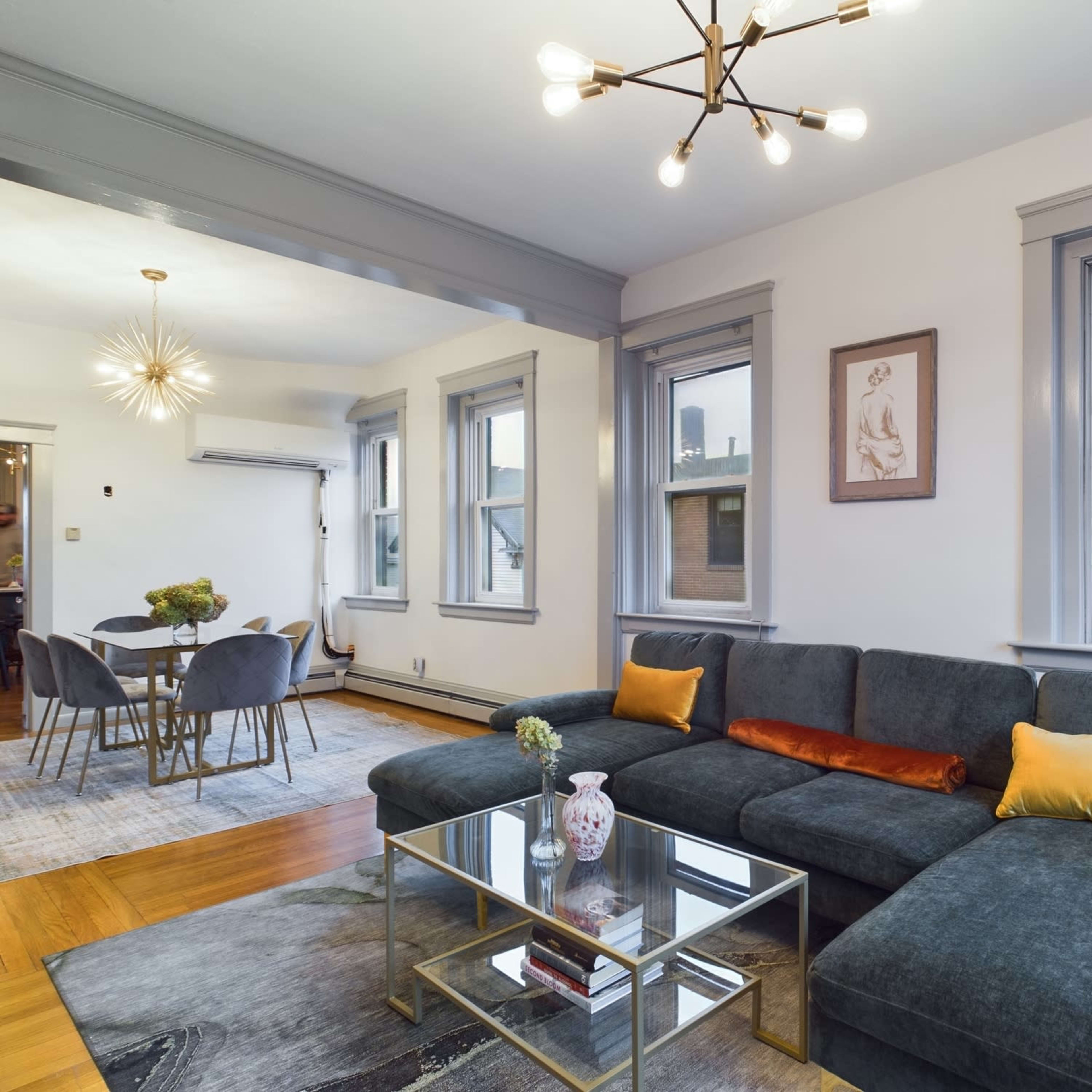 A modern living room with a dark gray sectional sofa, a glass coffee table, and two large windows, leading to a dining area with a round table and chairs.