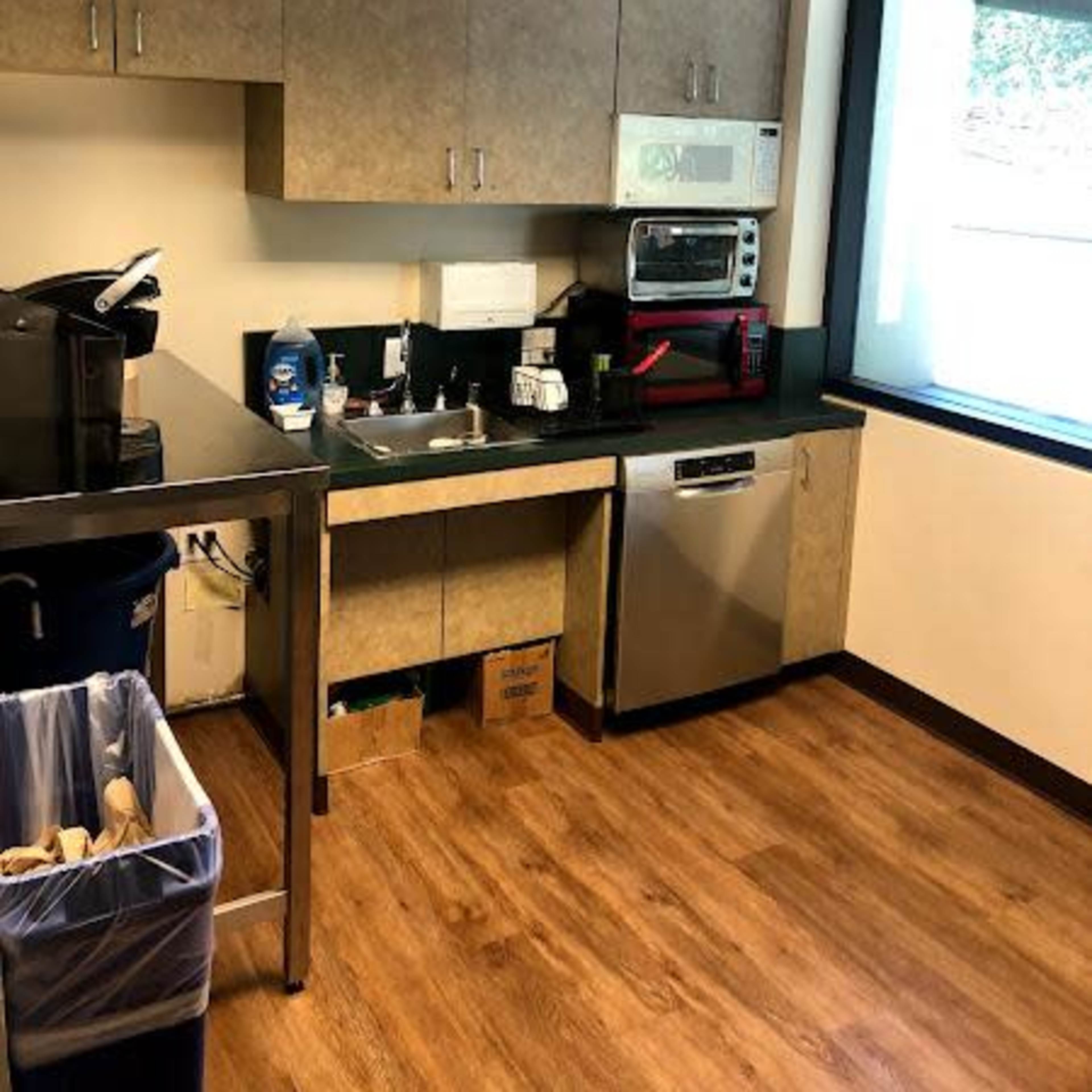 A kitchenette with cabinets, a sink, a microwave, a coffee maker, and a dishwasher, beside a trash bin.