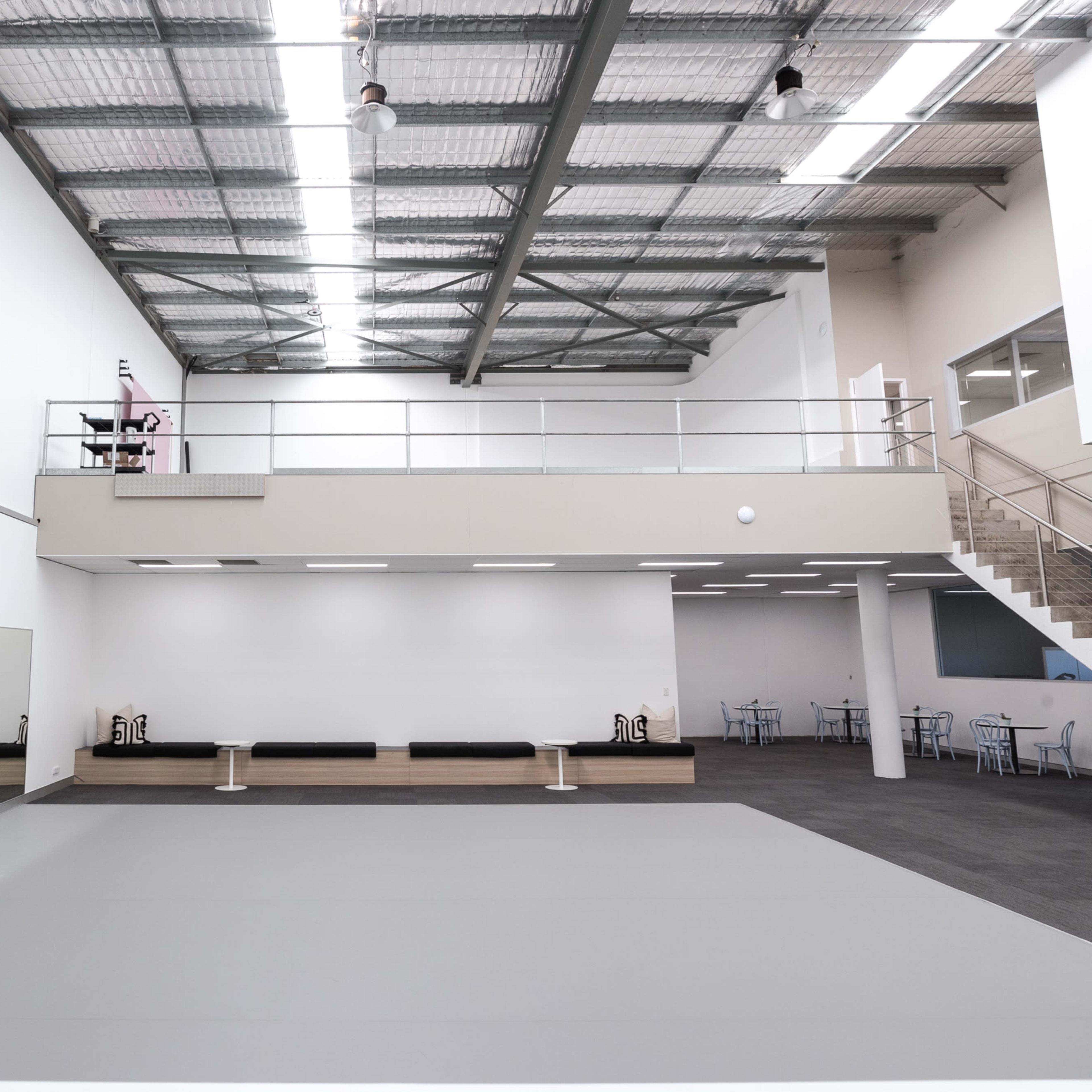 A spacious, brightly lit dance studio with a mirrored wall, a gray floor, and a mezzanine level overlooking the main area.