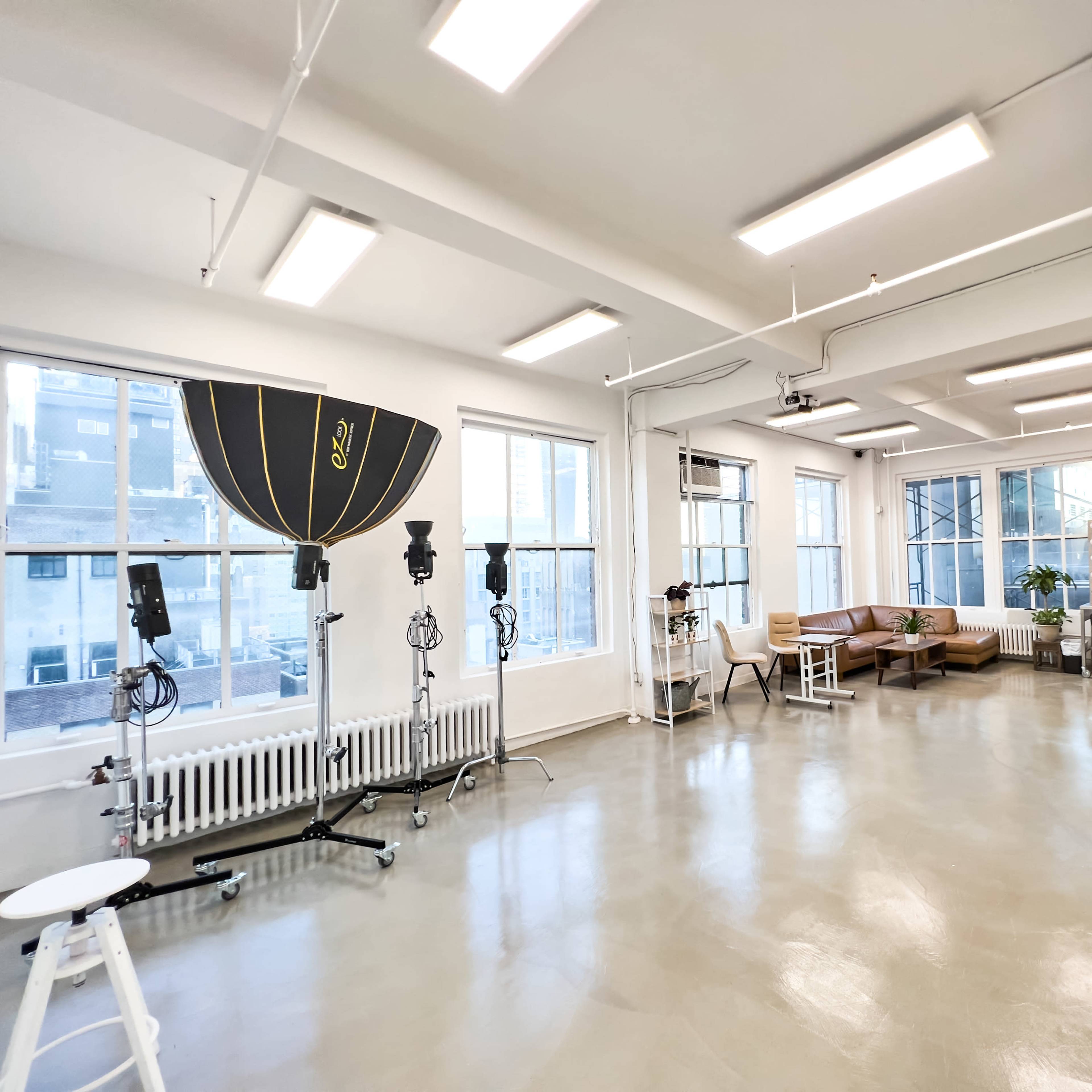A spacious, well-lit studio with large windows, featuring photography equipment, seating areas, and a clean floor.