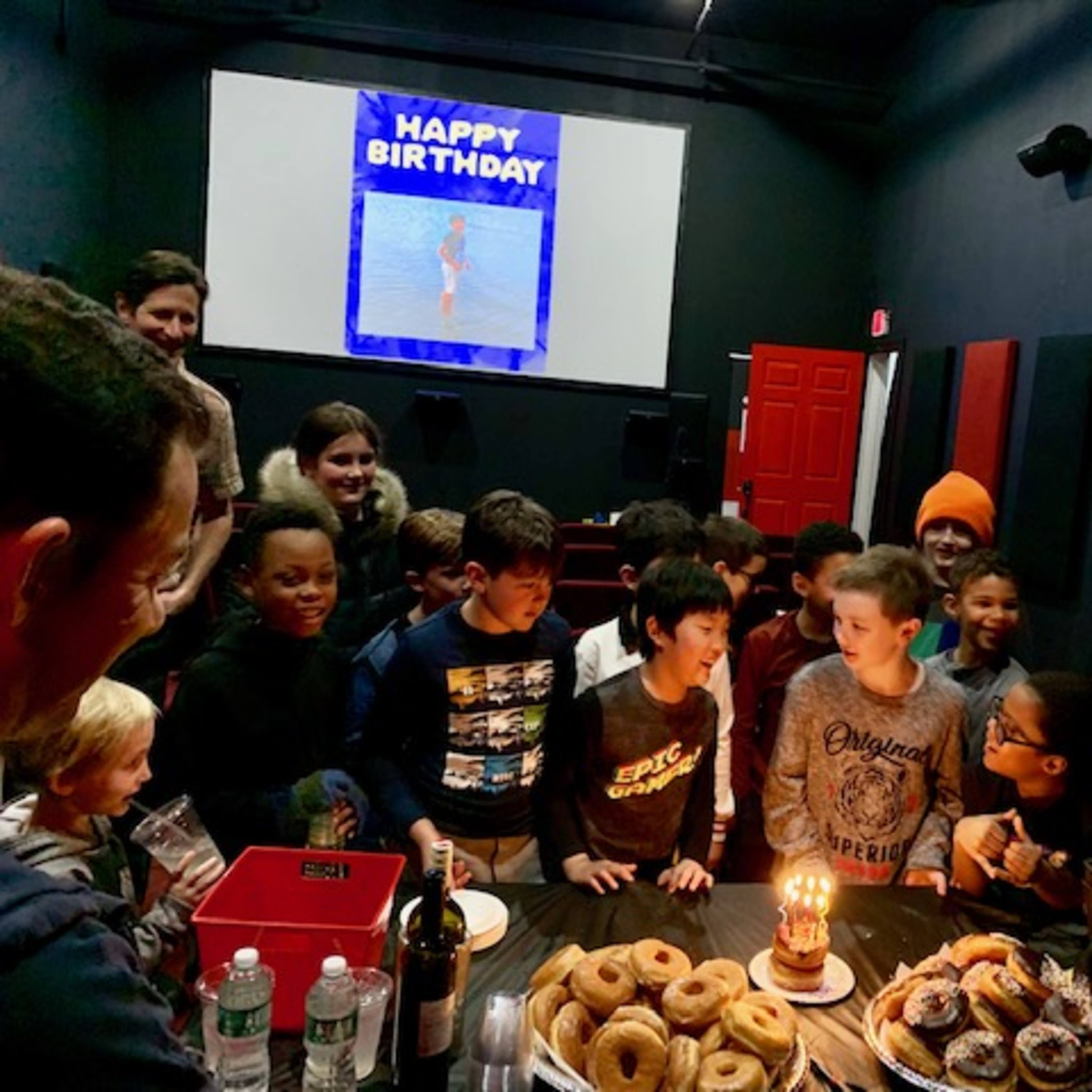 A group of children gathers around a table with donuts and a birthday cake, celebrating with a "Happy Birthday" sign displayed on a screen in the background.