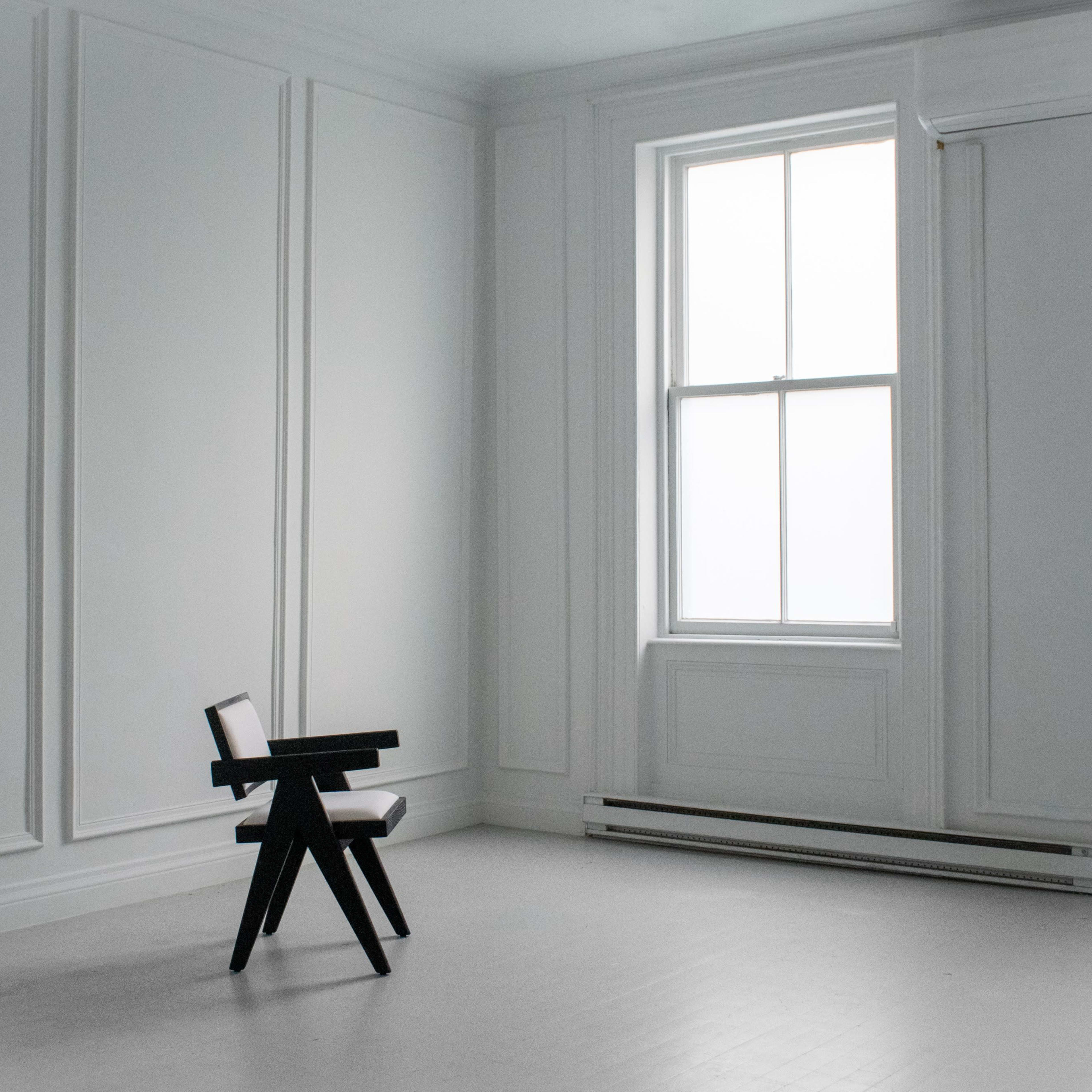 A minimalist room with white walls, a large window, and a single black chair located in the center.