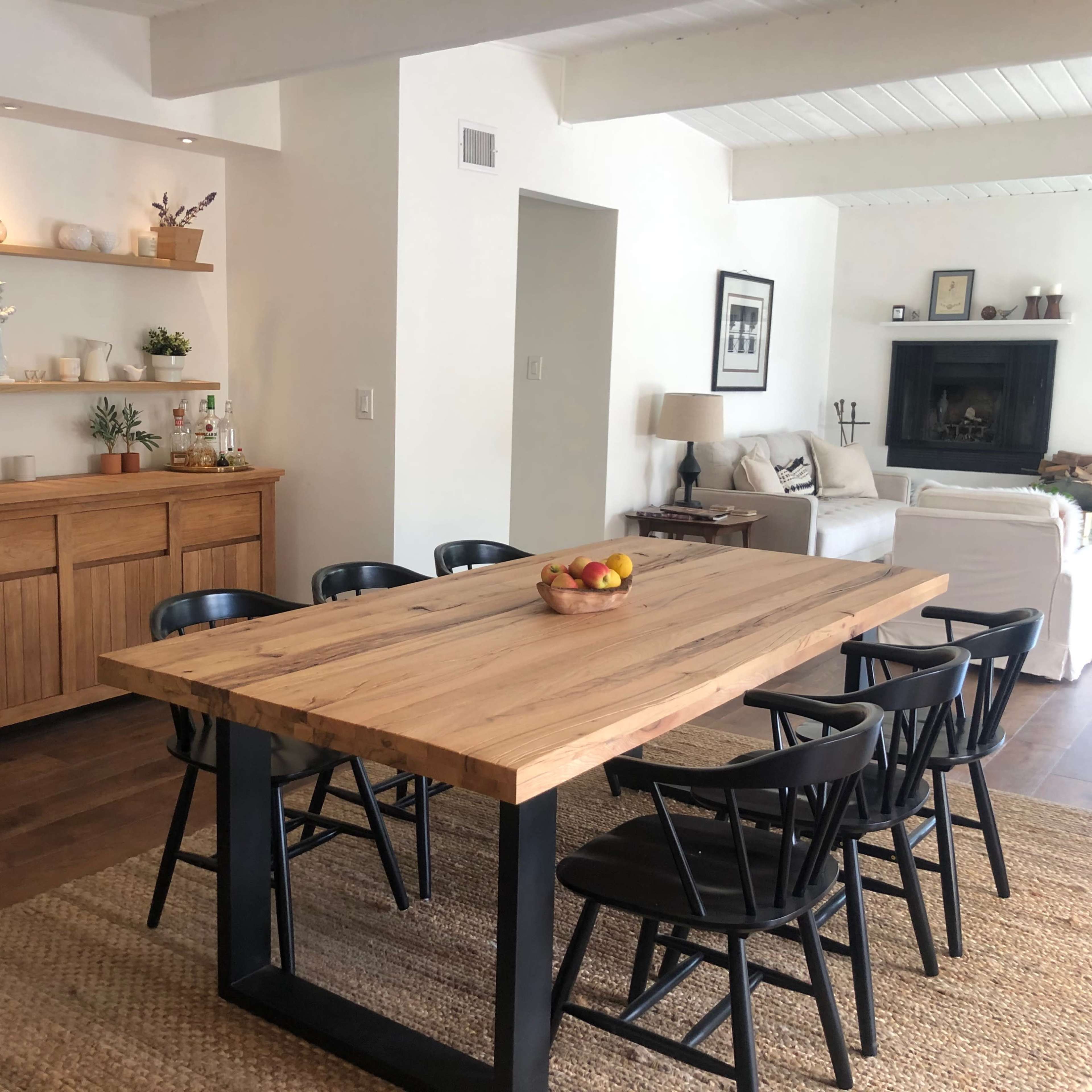 A wooden dining table with black chairs is positioned in a room featuring a sideboard and shelves displaying decorative items.