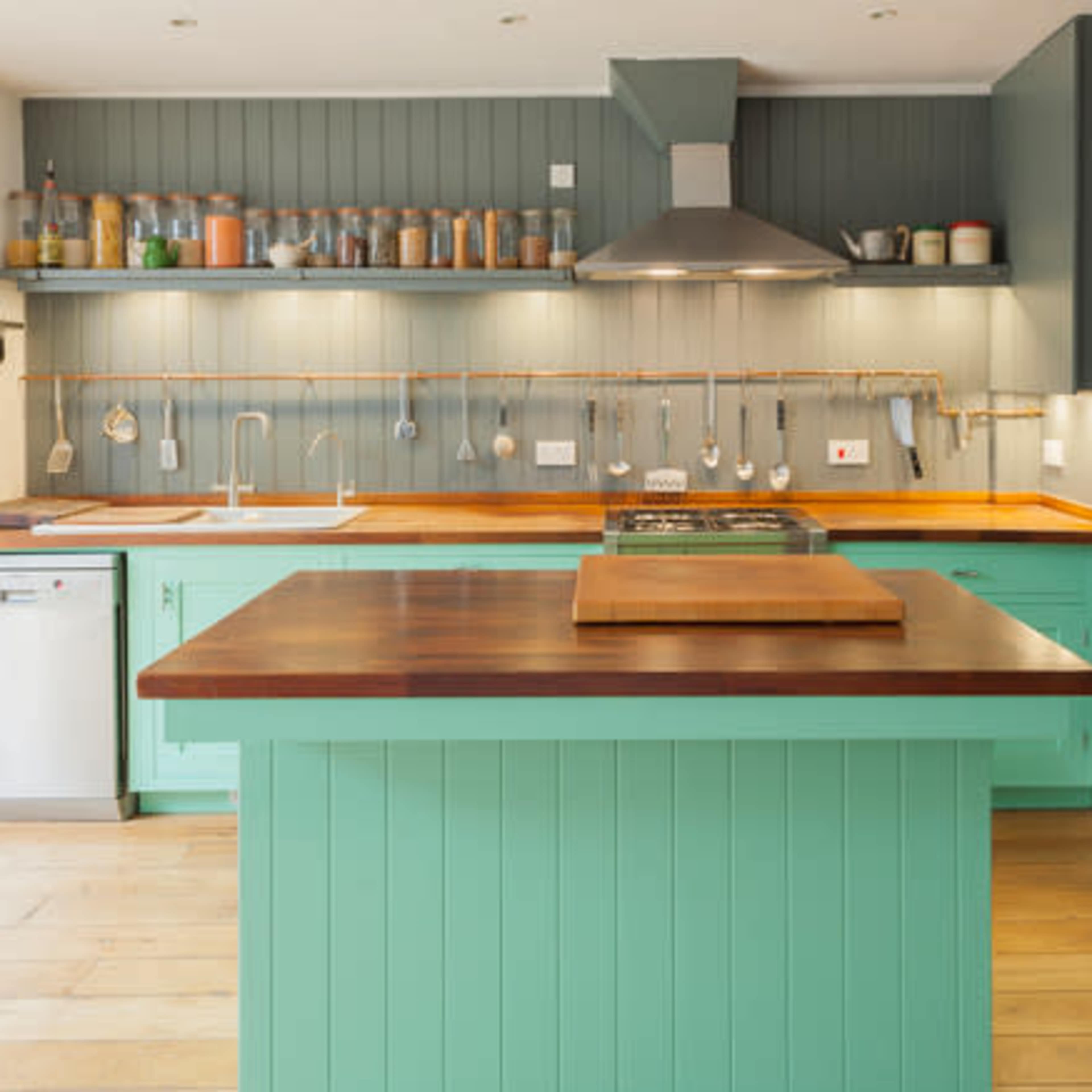 A modern kitchen with teal cabinetry, wooden countertops, and stainless steel appliances, featuring an island in the center.