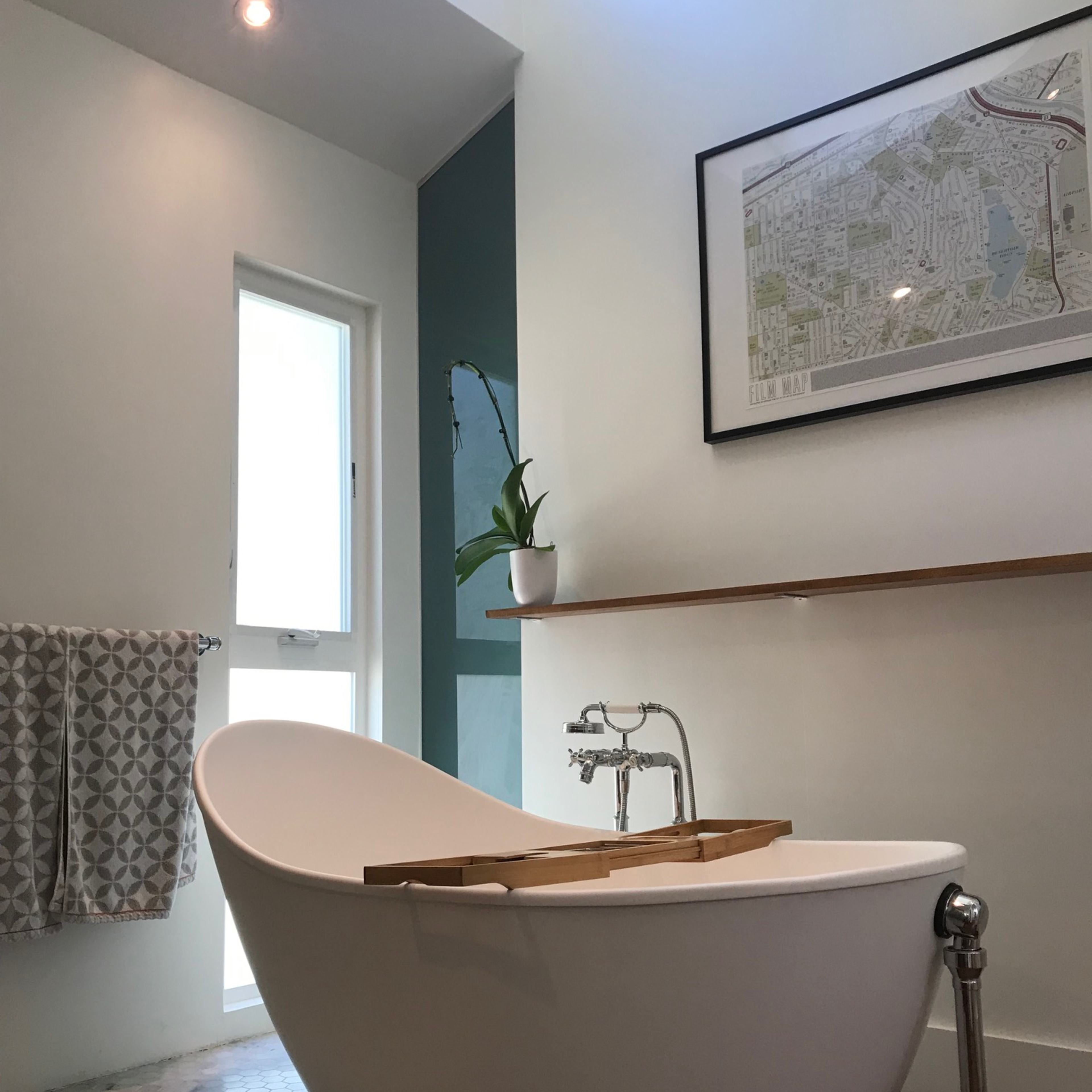 A modern bathroom featuring a freestanding white bathtub, a wooden shelf, a map on the wall, and a window allowing natural light.