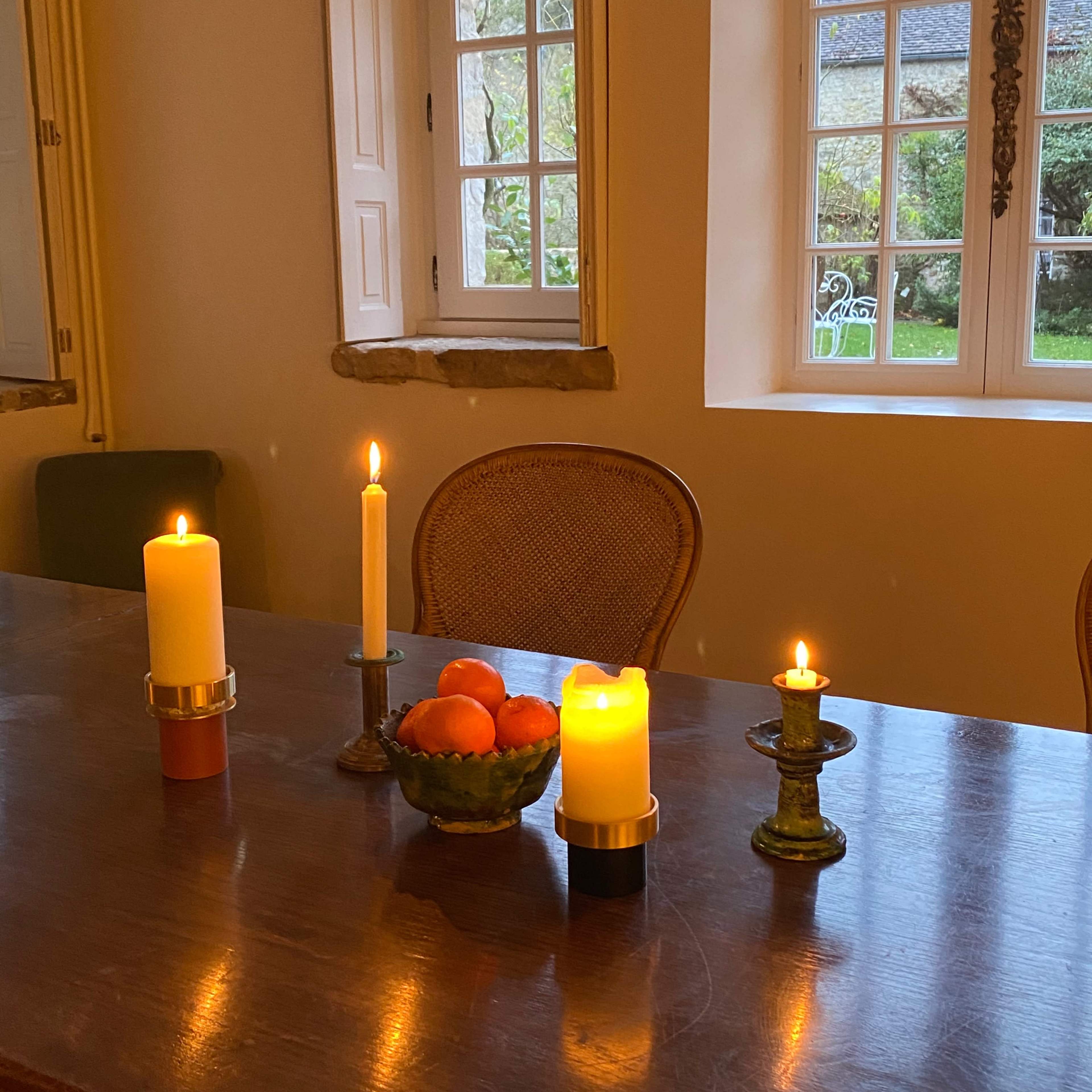 A wooden table is set with three candles and a bowl of oranges, in a well-lit room with windows overlooking a garden.