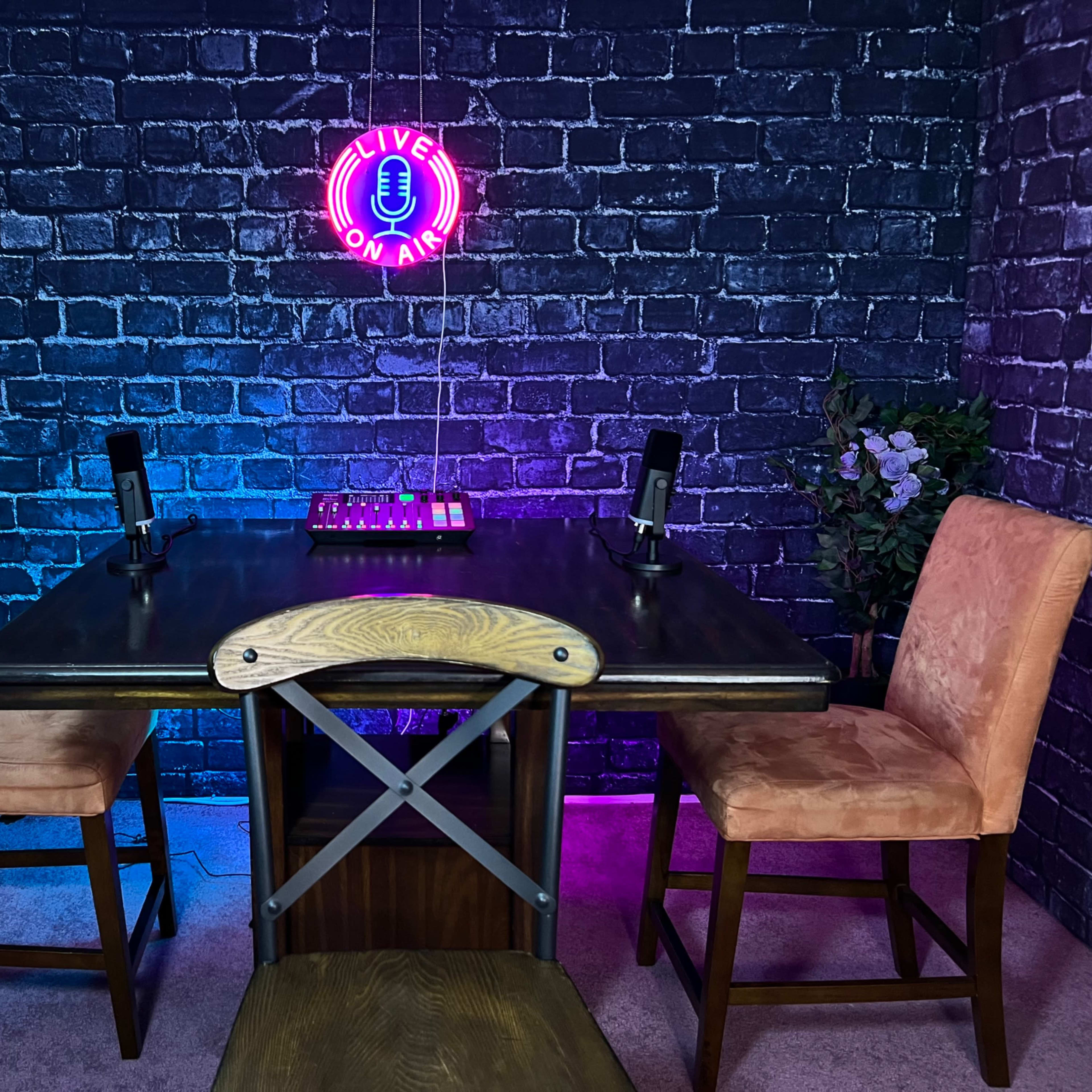 A dark brick wall serves as the backdrop for a small table with two microphones and two chairs, illuminated by a neon "LIVE ON AIR" sign.