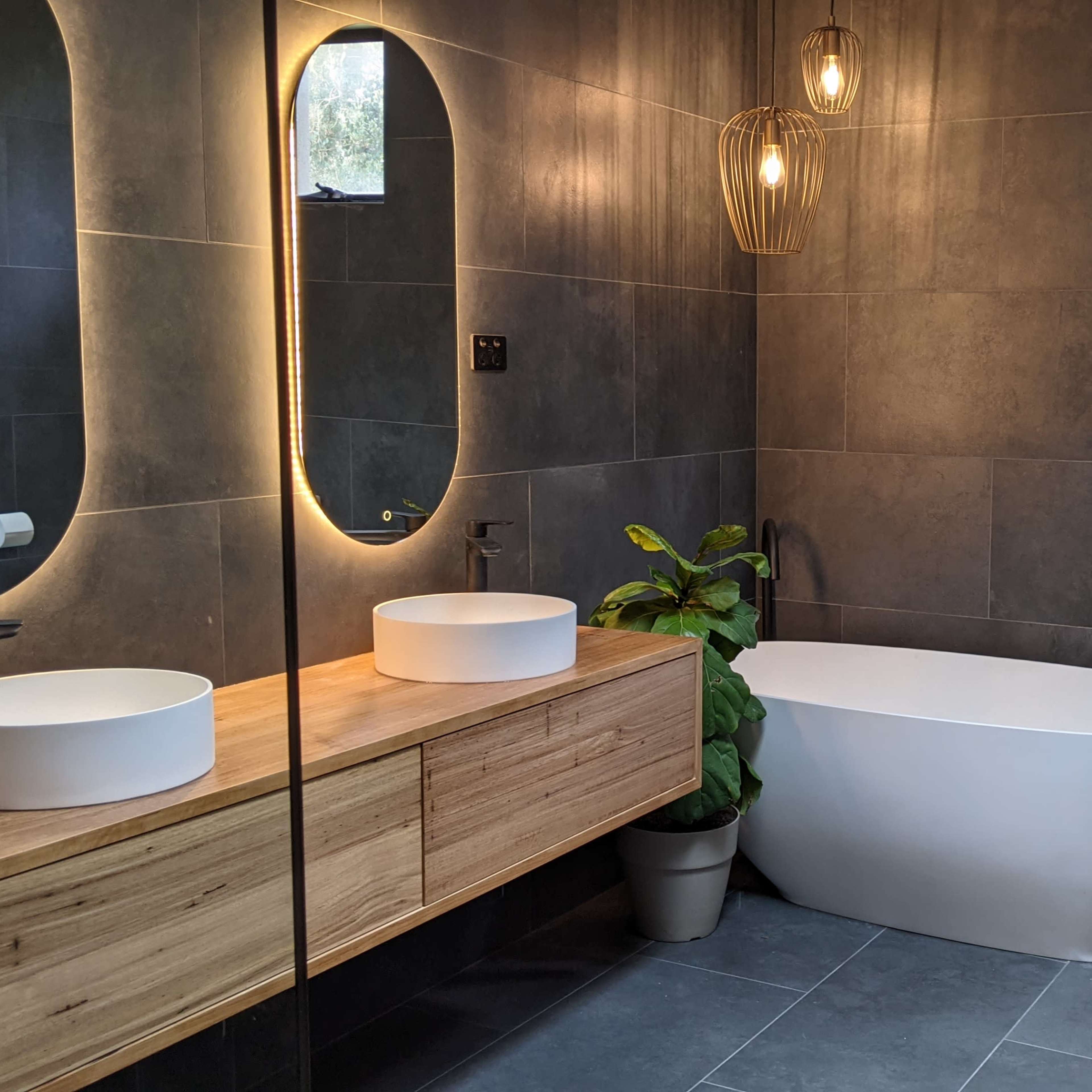 A modern bathroom features a freestanding bathtub, double sinks on a wooden vanity, and a wall-mounted toilet, all set against dark tiled walls.