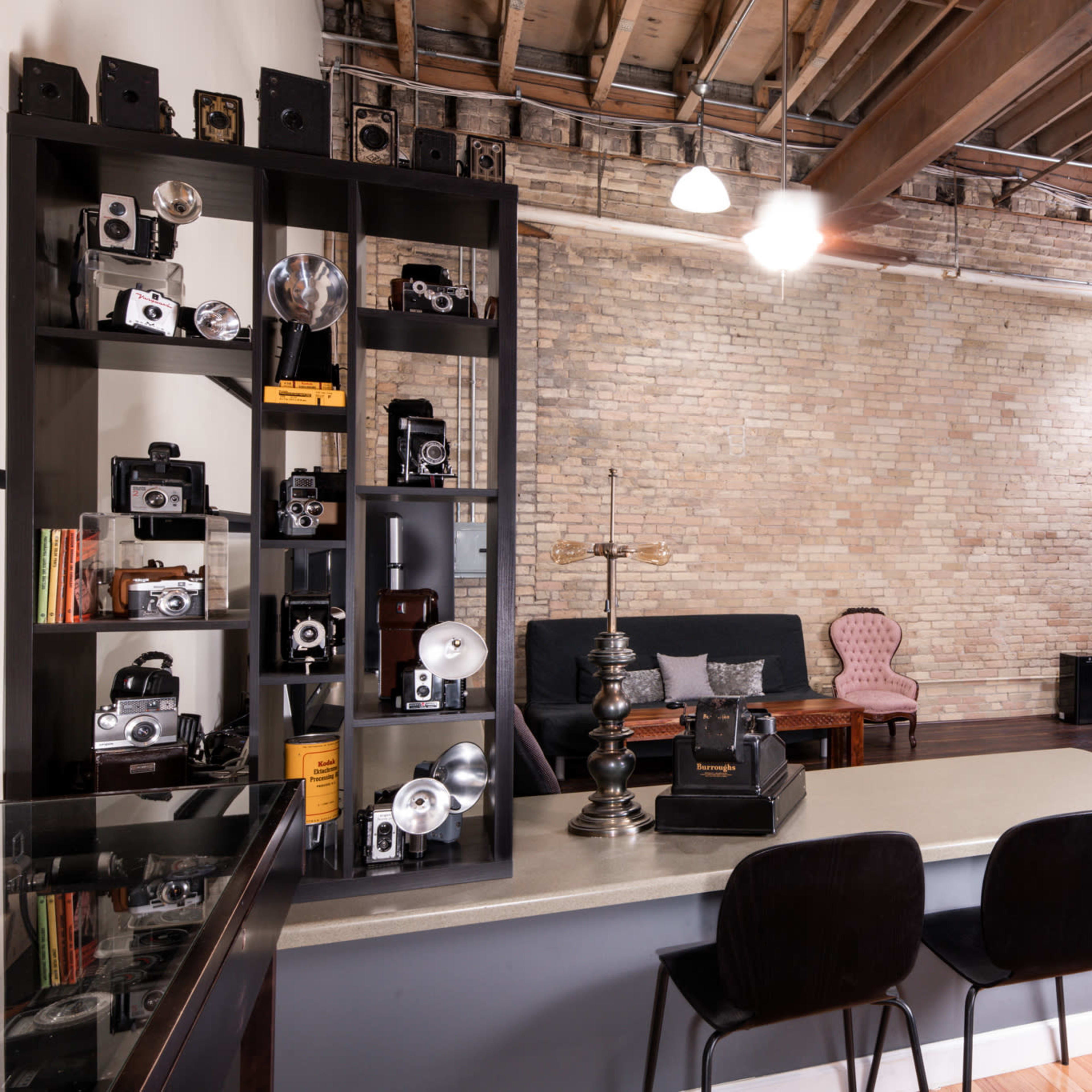 A spacious photography studio with a brick wall, decorative cameras on a black shelf, a seating area with a vintage chair, and large lighting equipment in the background.