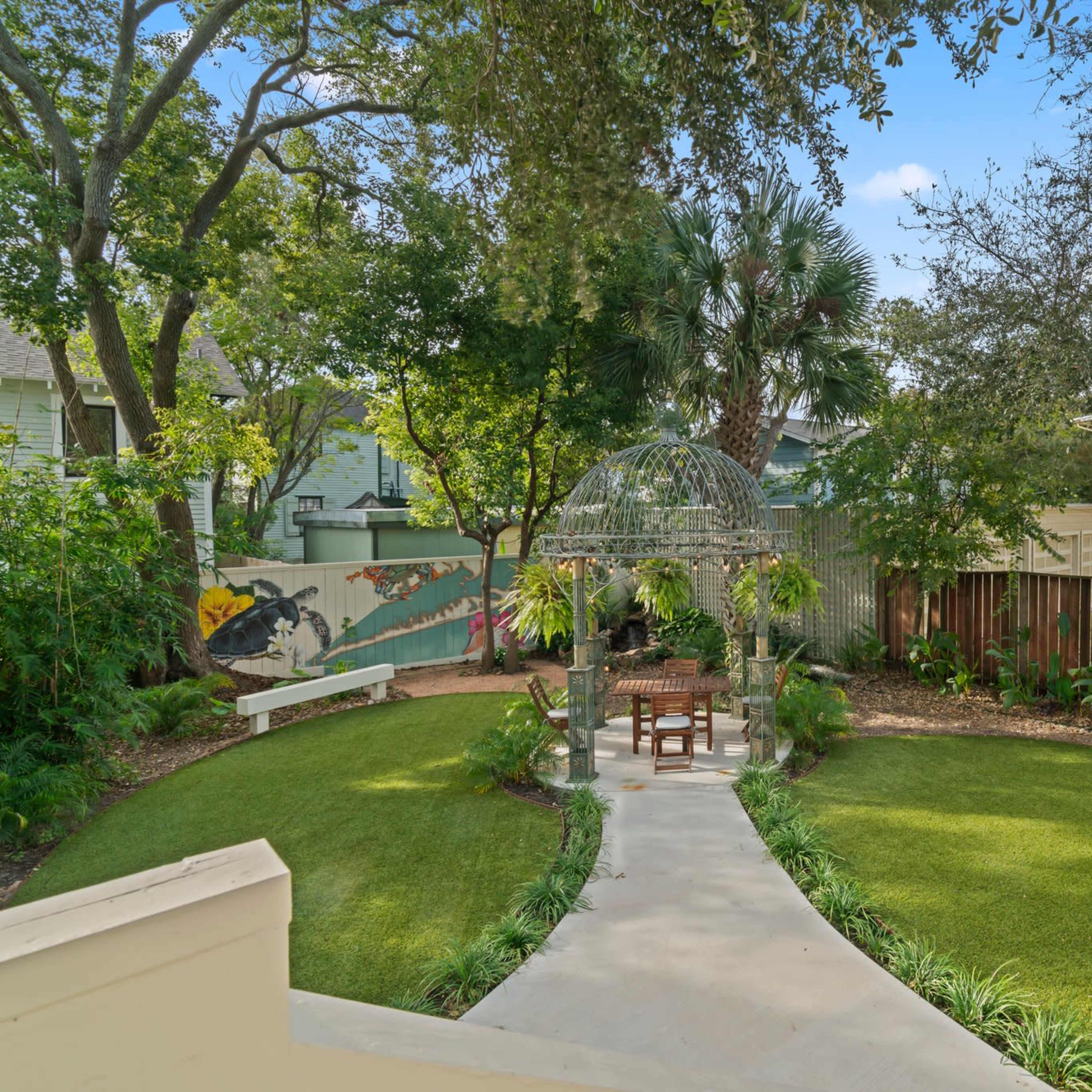 A neatly landscaped garden featuring a circular path, a gazebo, a wooden chair, and various plants surrounded by trees and a wooden fence.