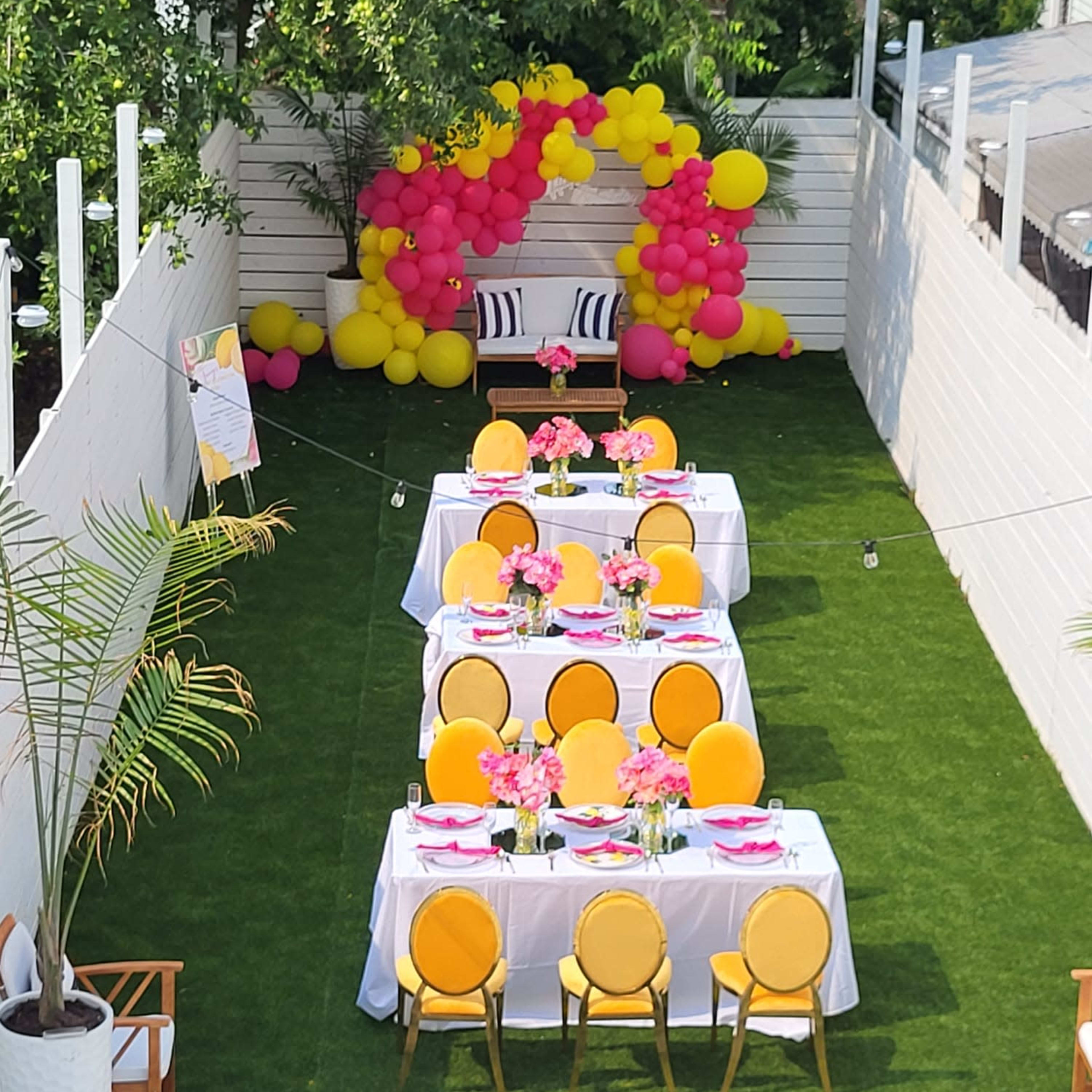 An outdoor party setup with two long tables decorated with flowers and bright table settings, surrounded by colorful balloons and greenery.