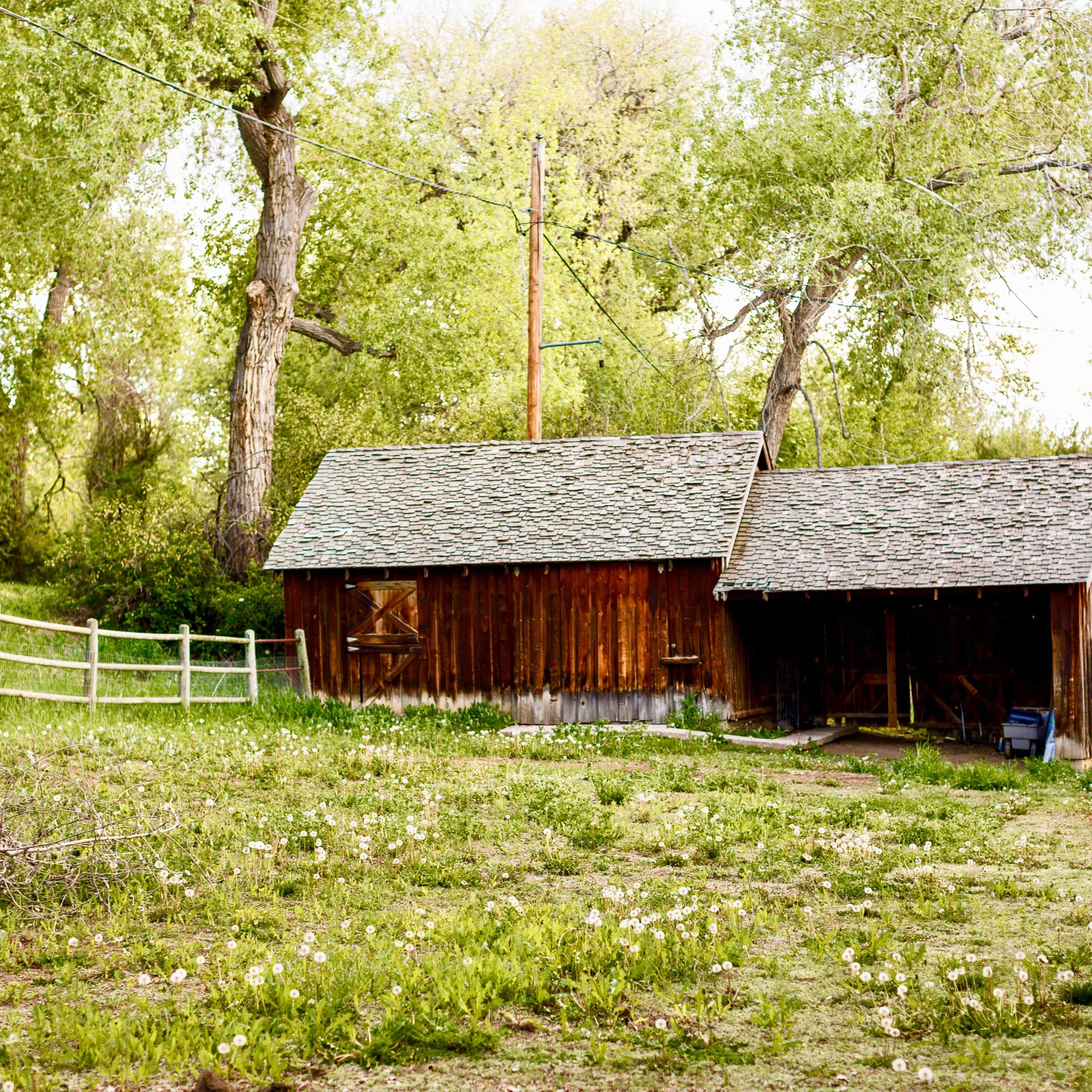 A wooden barn sits on a grassy area surrounded by trees and a fence.