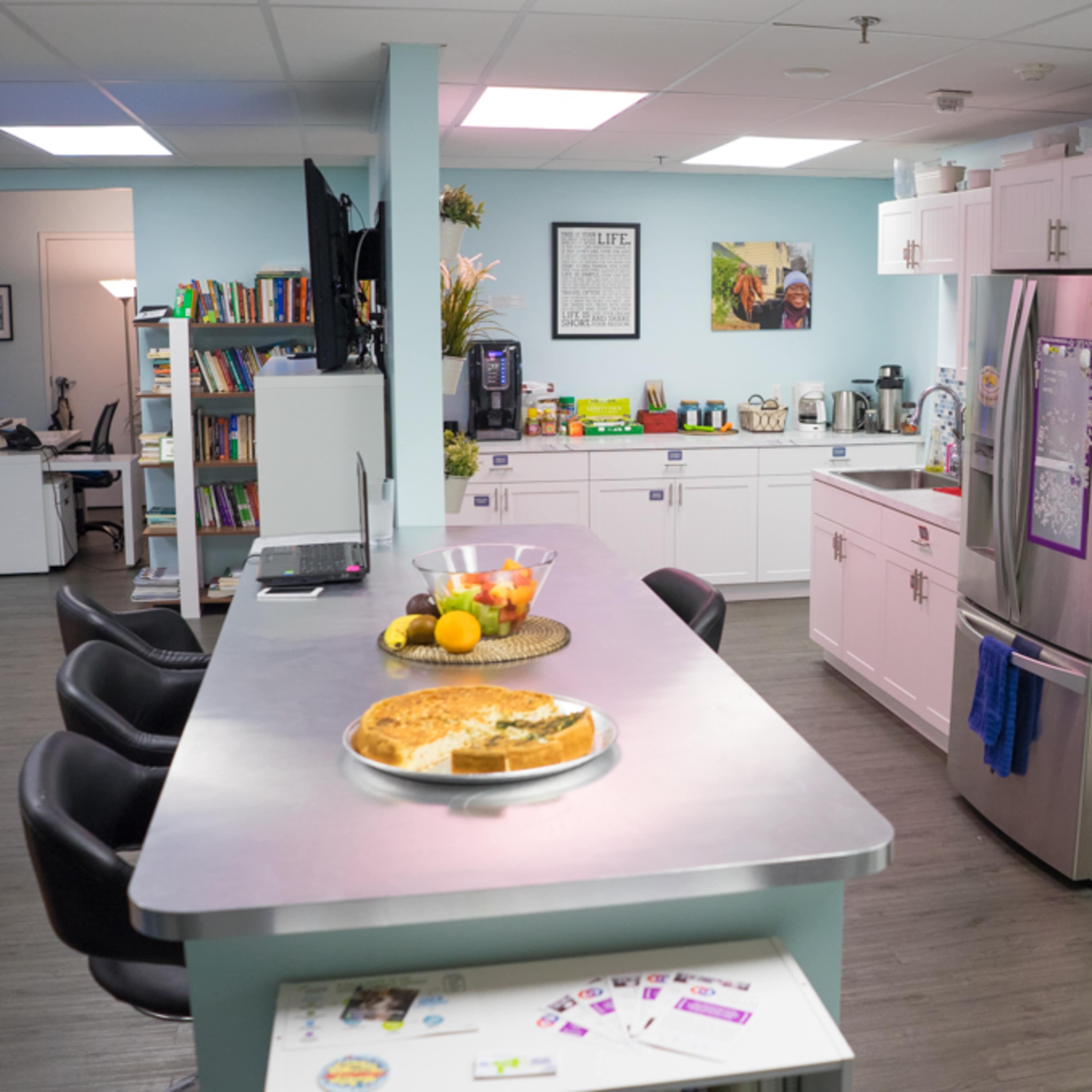 A modern kitchen with a large countertop, stainless steel appliances, and a fruit bowl, adjacent to a cozy workspace and bookshelves.