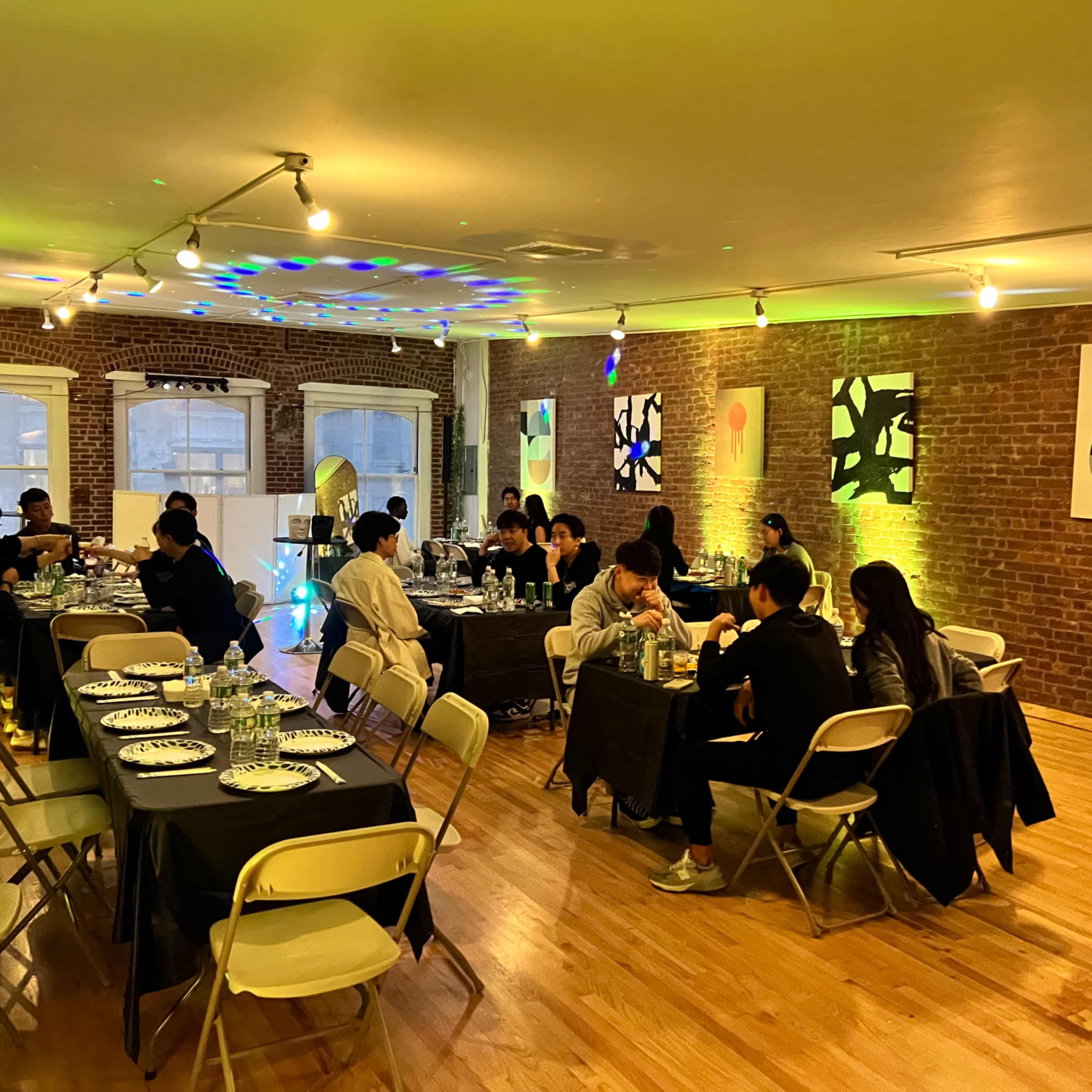 A group of people explore an art gallery featuring various paintings and sculptures displayed on pedestals in a room with wooden flooring and brick walls.