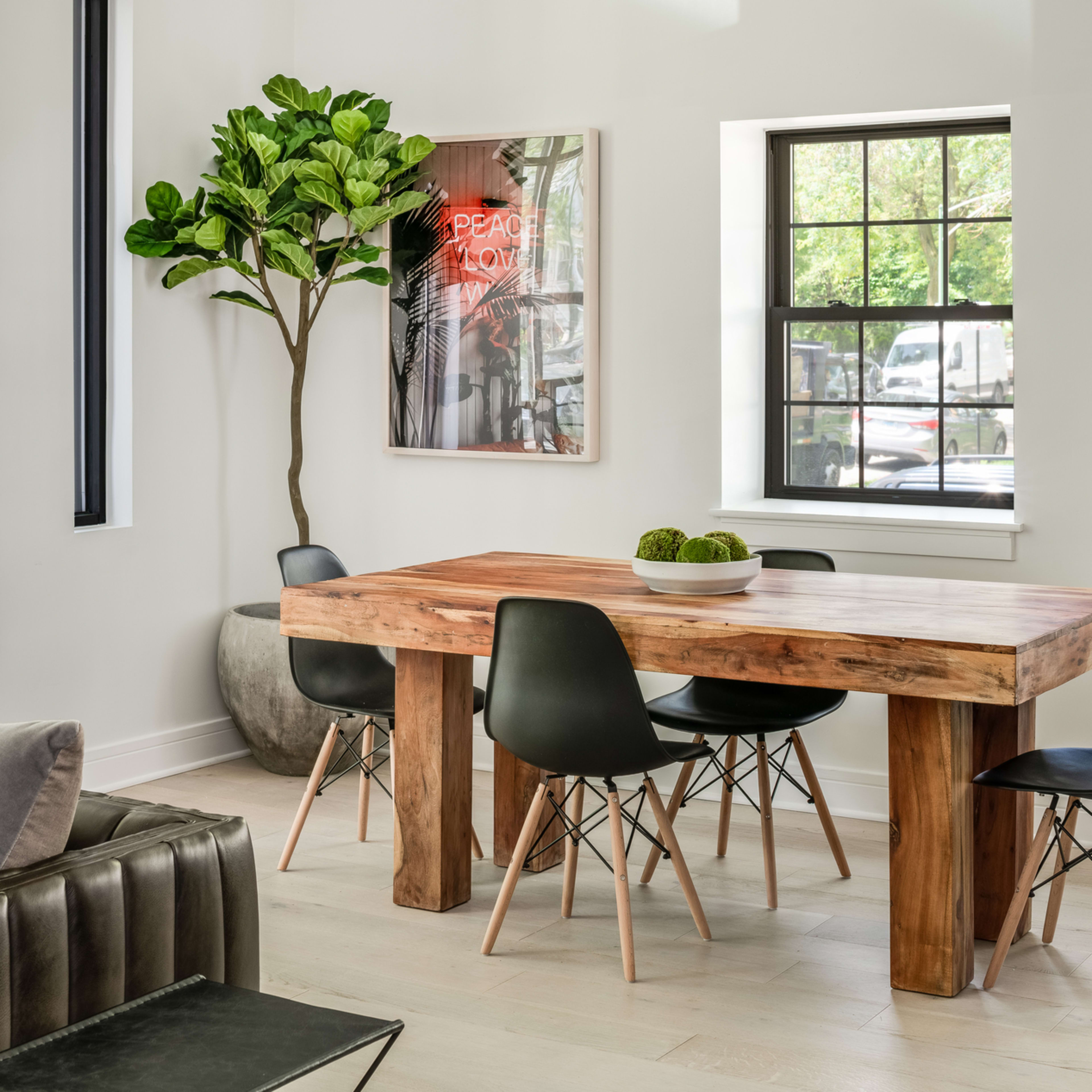 A wooden dining table surrounded by black chairs is positioned near a window and a decorative plant in a modern interior.