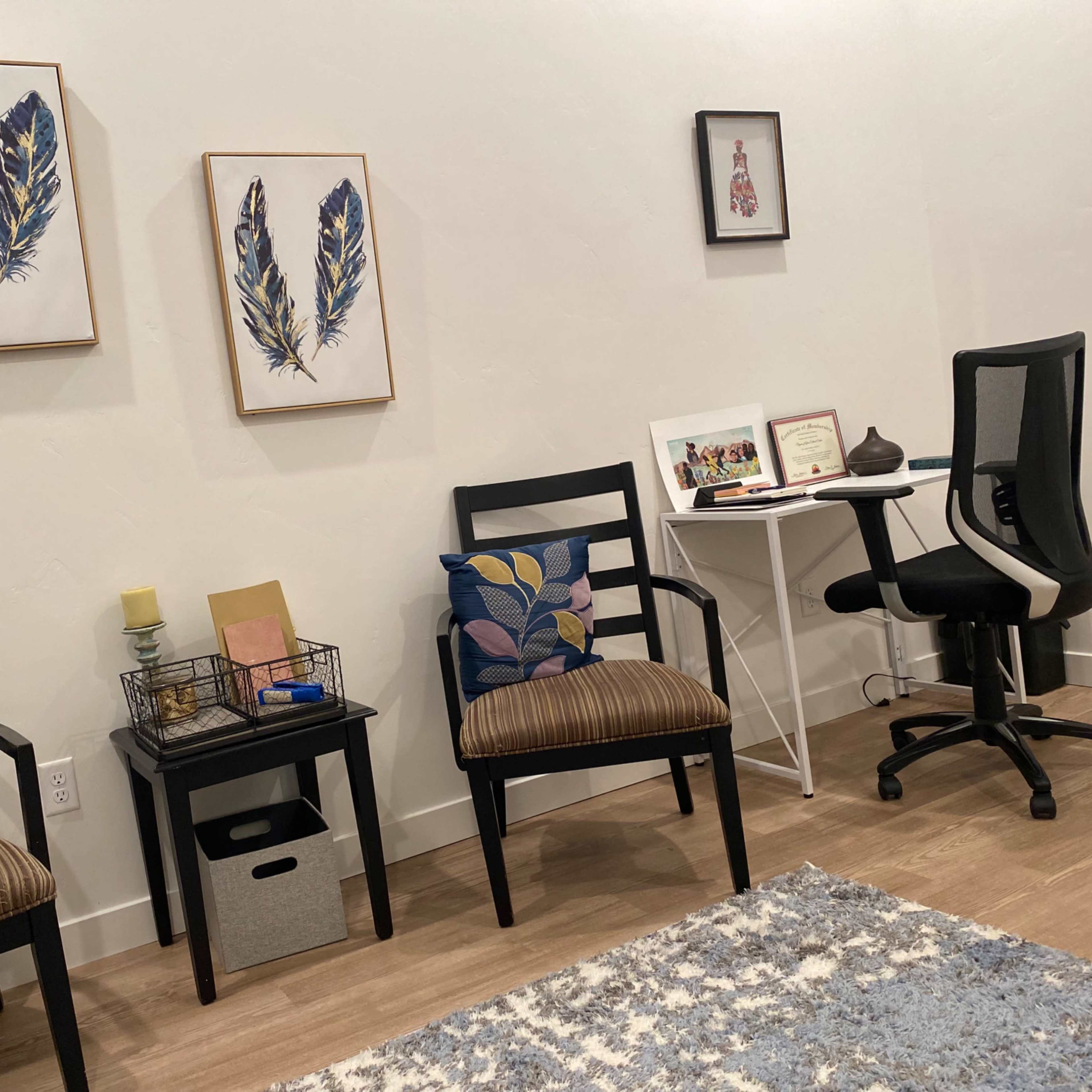 A waiting area with two black chairs, decorative feather art on the wall, a small side table with a candle, and a desk with a chair in the corner.
