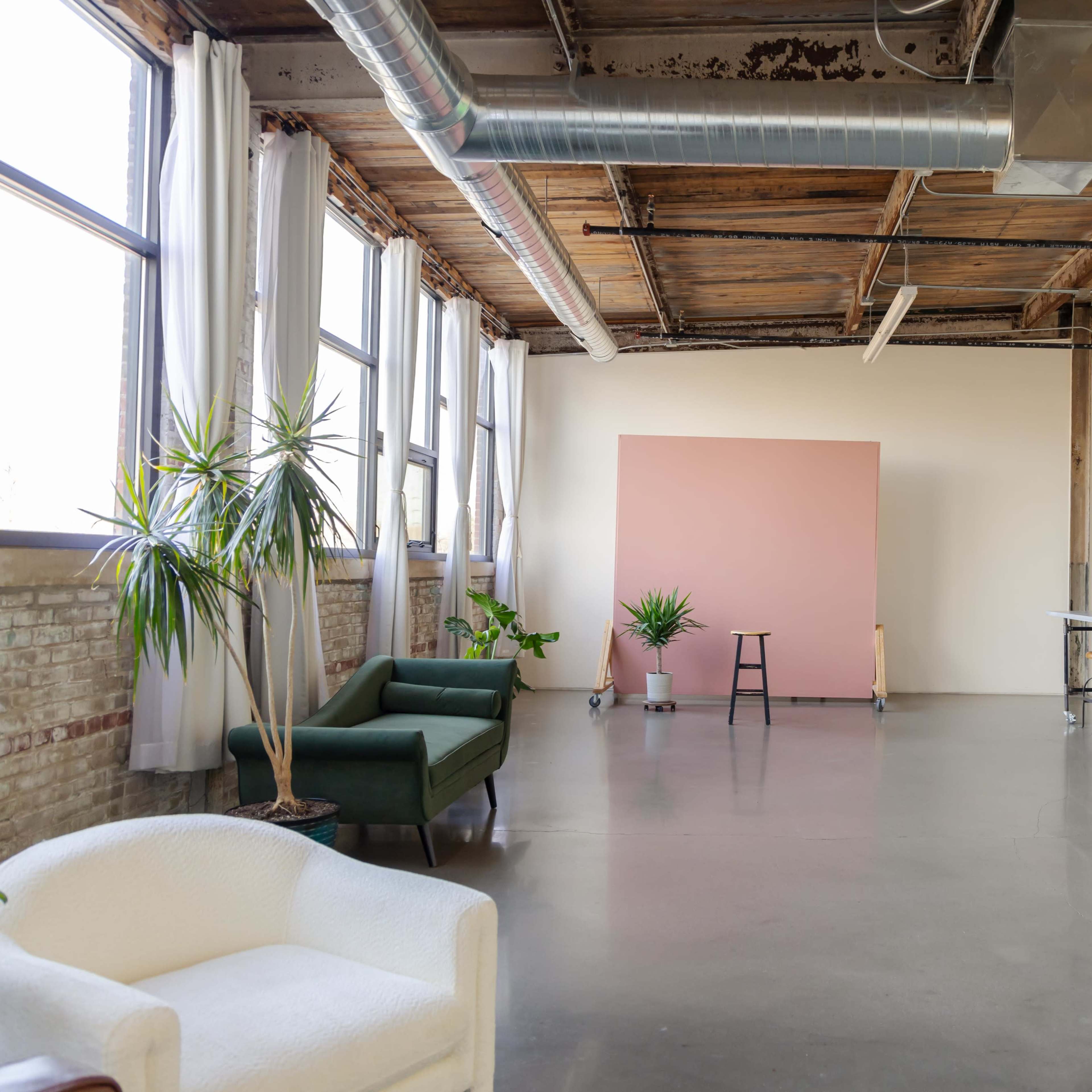 A spacious, industrial-style room with large windows, concrete floors, and various plants, featuring a pink backdrop and kitchen appliances along one wall.