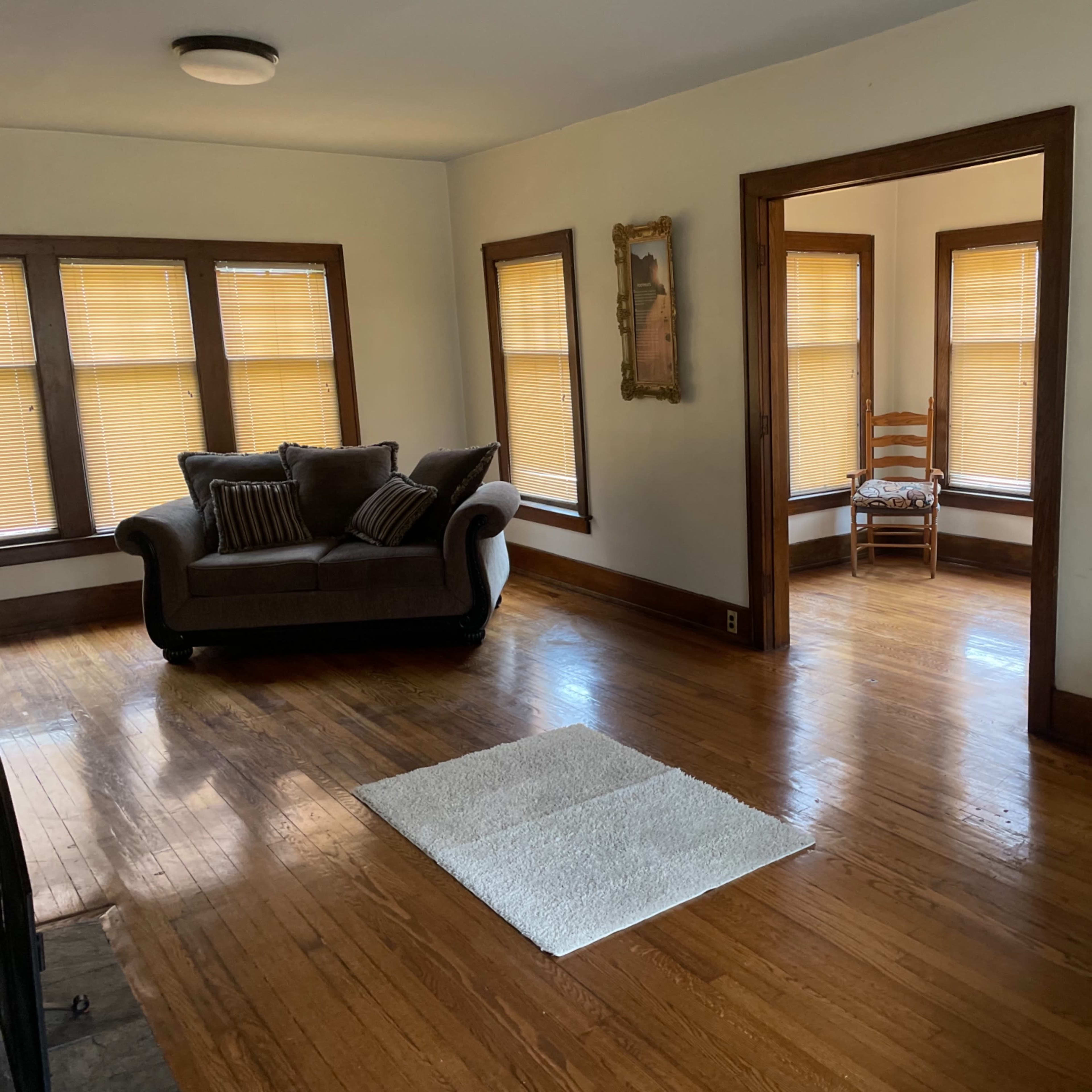 A living room features two sofas, a coffee table on a rug, and large windows with shades, leading to an adjacent room with a chair.