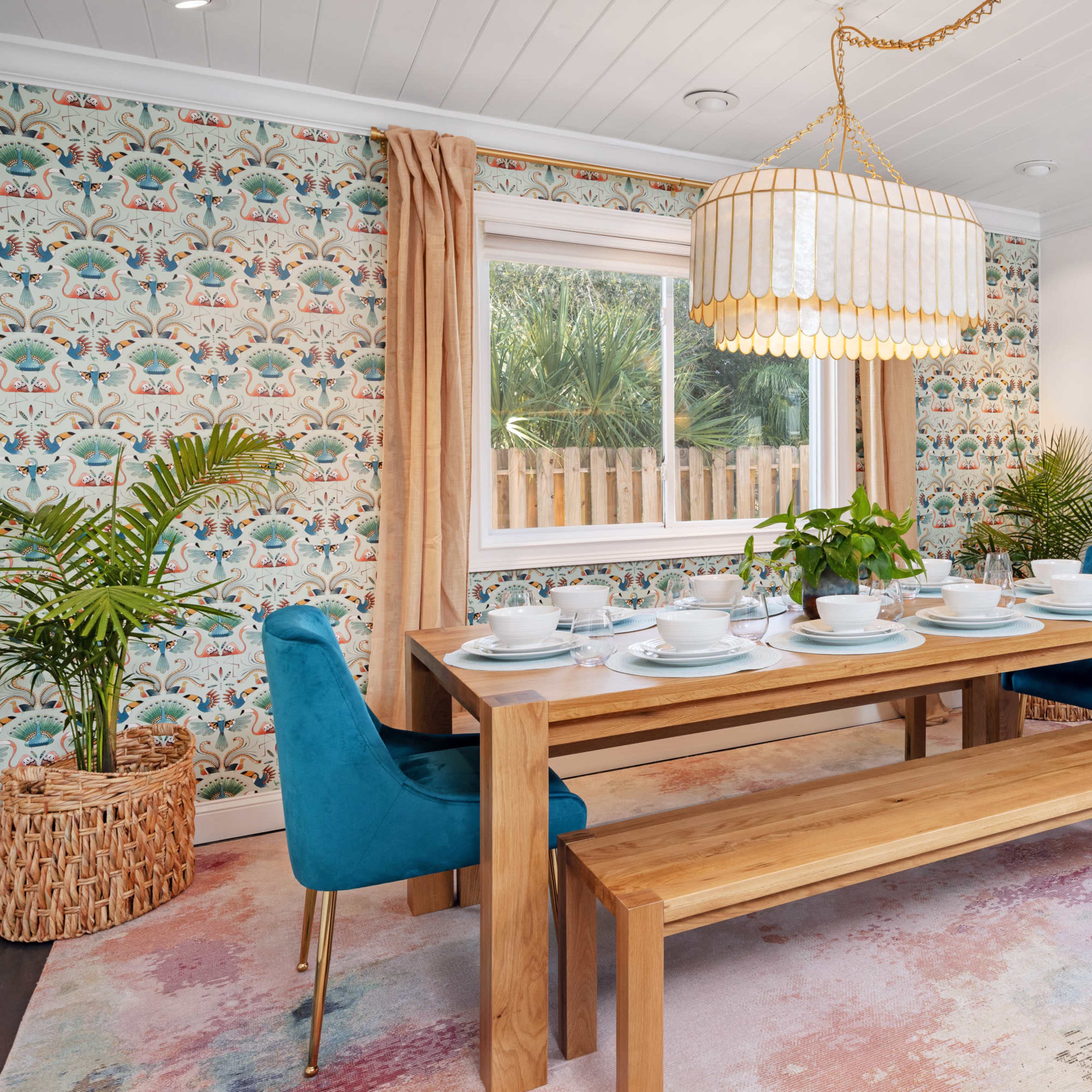 A dining room features a wooden table set for six, surrounded by blue chairs, a long chandelier above, and decorative wallpaper with vibrant patterns.